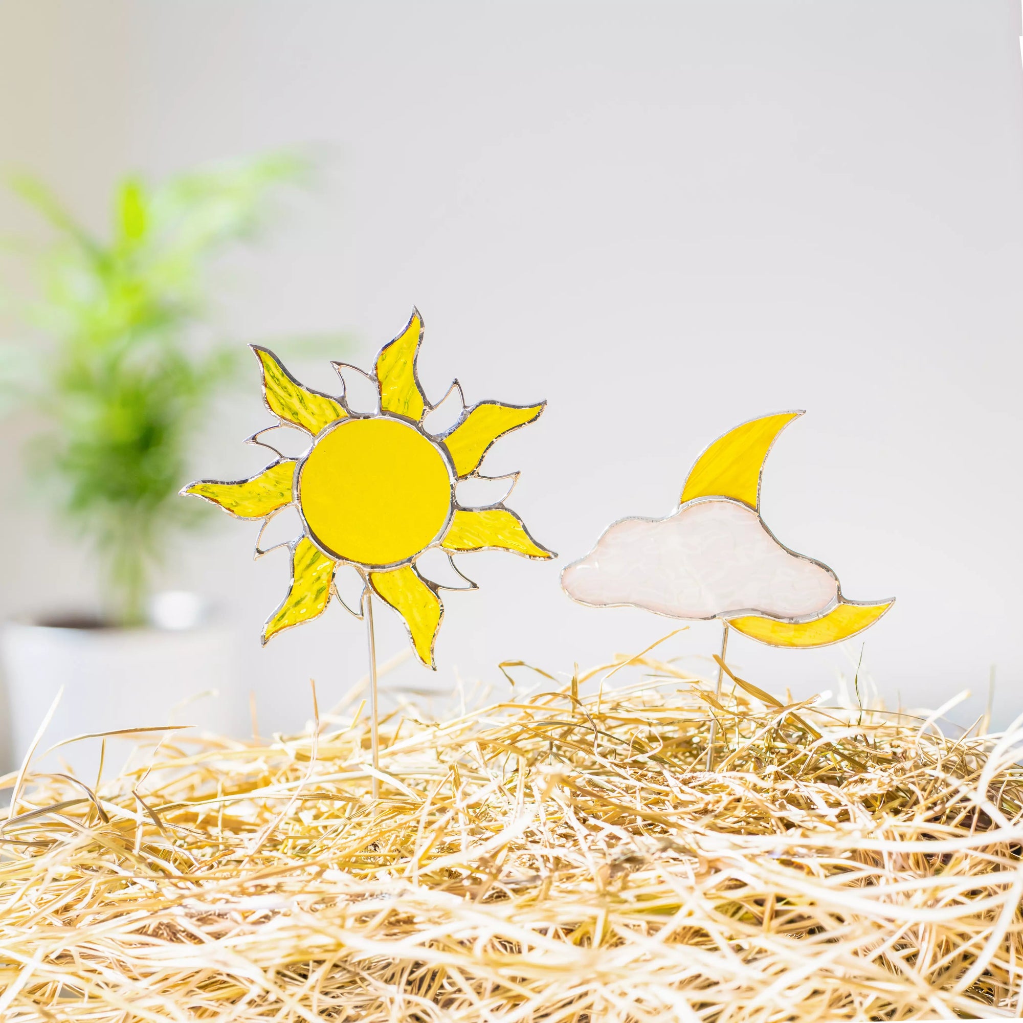 Stained glass sun and moon plant stakes on a bed of straw with a blurred indoor background.