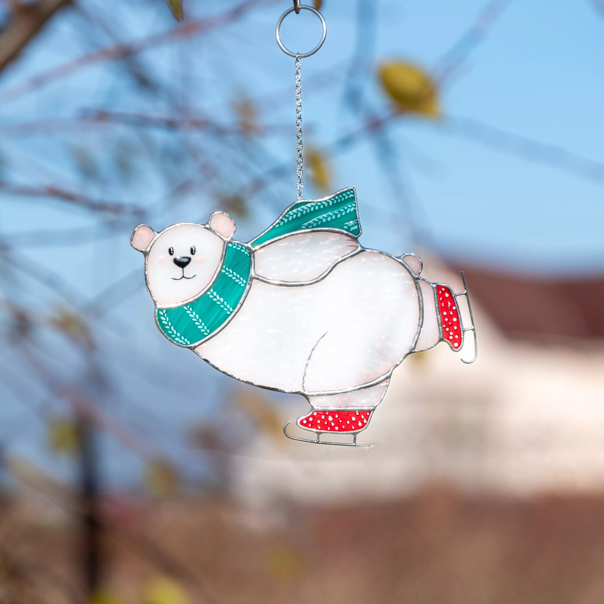 Stained glass suncatcher featuring a polar bear with a green scarf and red skates against a blurred natural background.