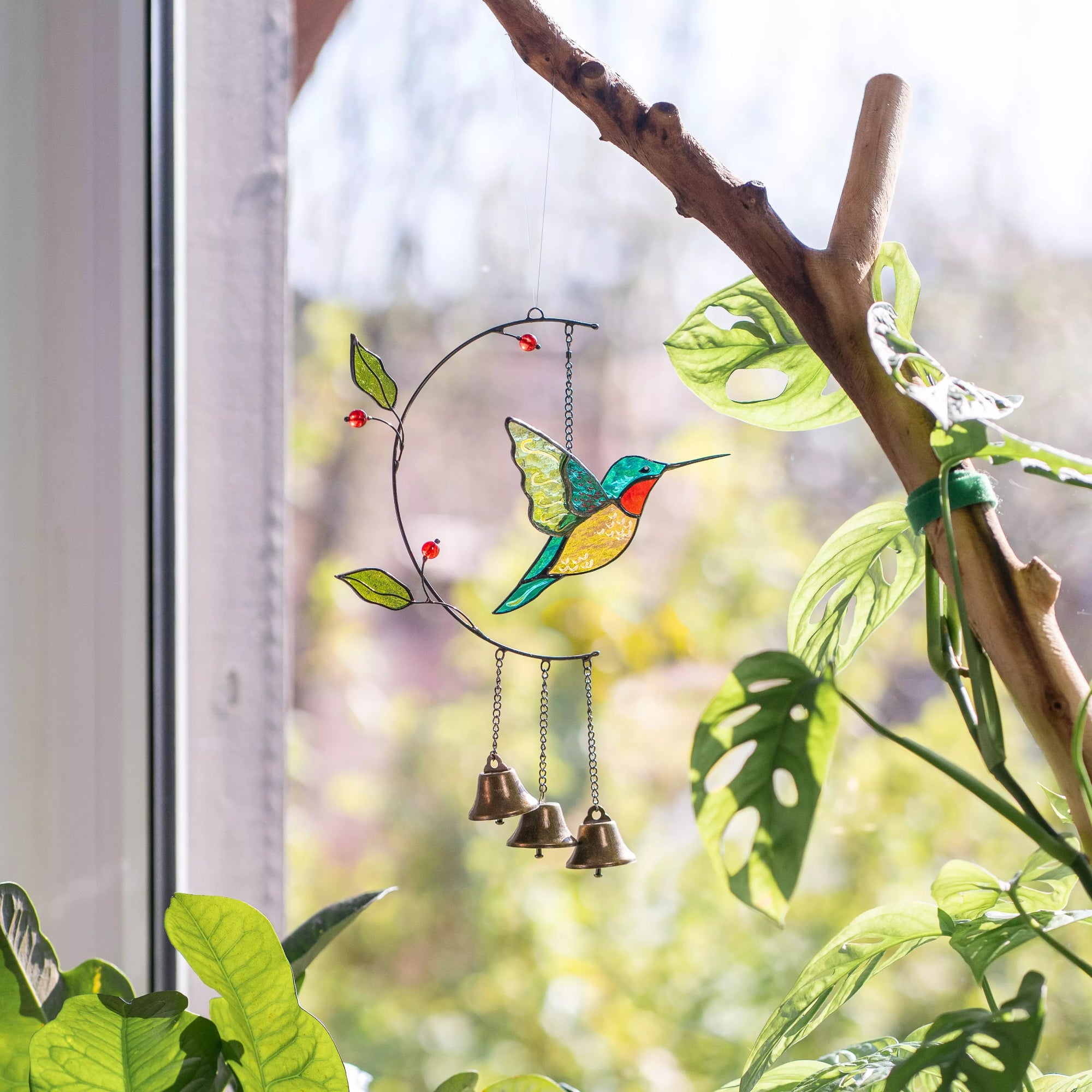 Stained glass wind chime shaped like a hummingbird hanging on a branch with green leaves.