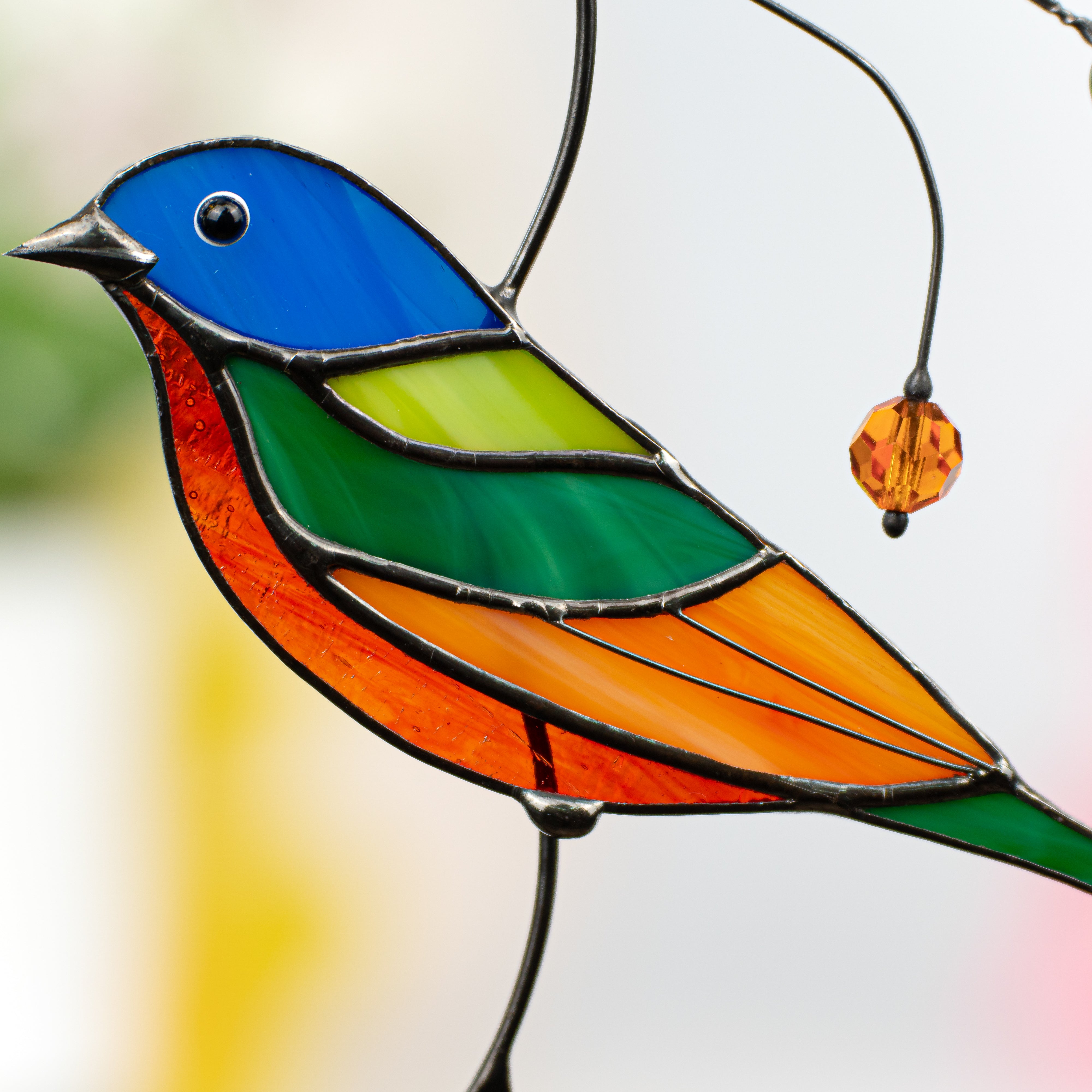 Close-up of bunting bird’s blue, green, and orange stained glass body with a hanging amber bead.