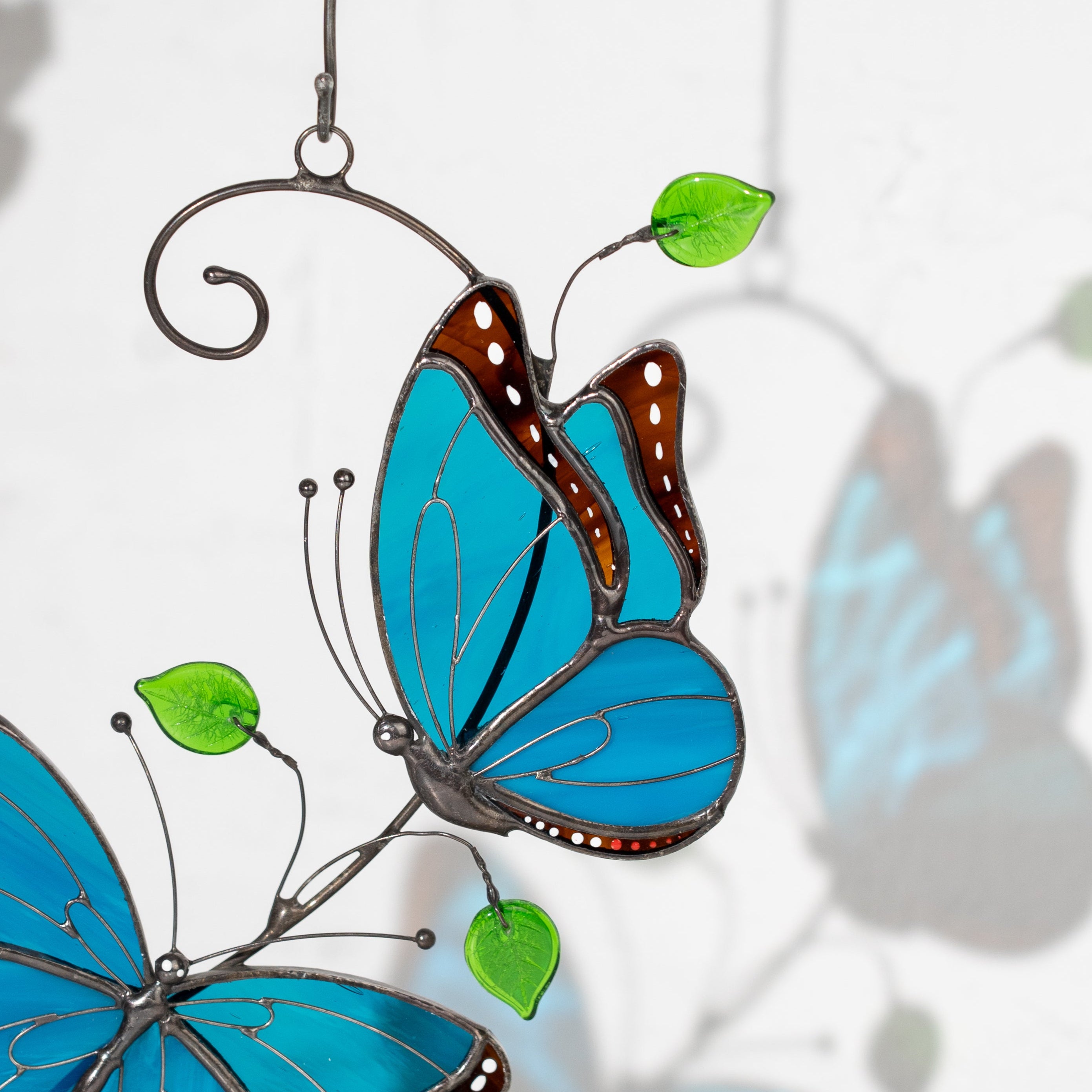Close-up of two blue stained glass morpho butterflies with brown borders and green leaves, hanging as a suncatcher.
