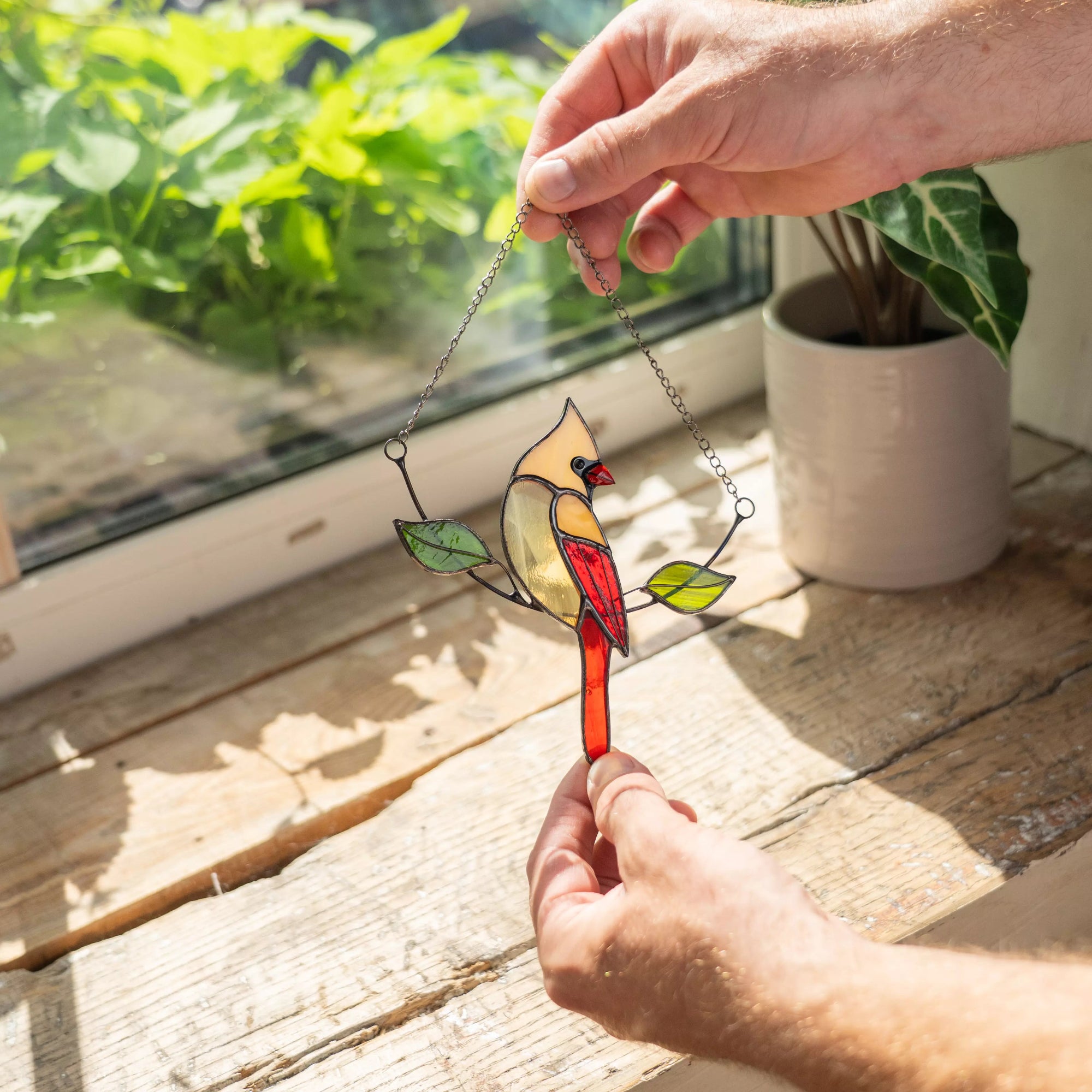 Stained glass female cardinal on a chain held by a hand with a window and plant in the background