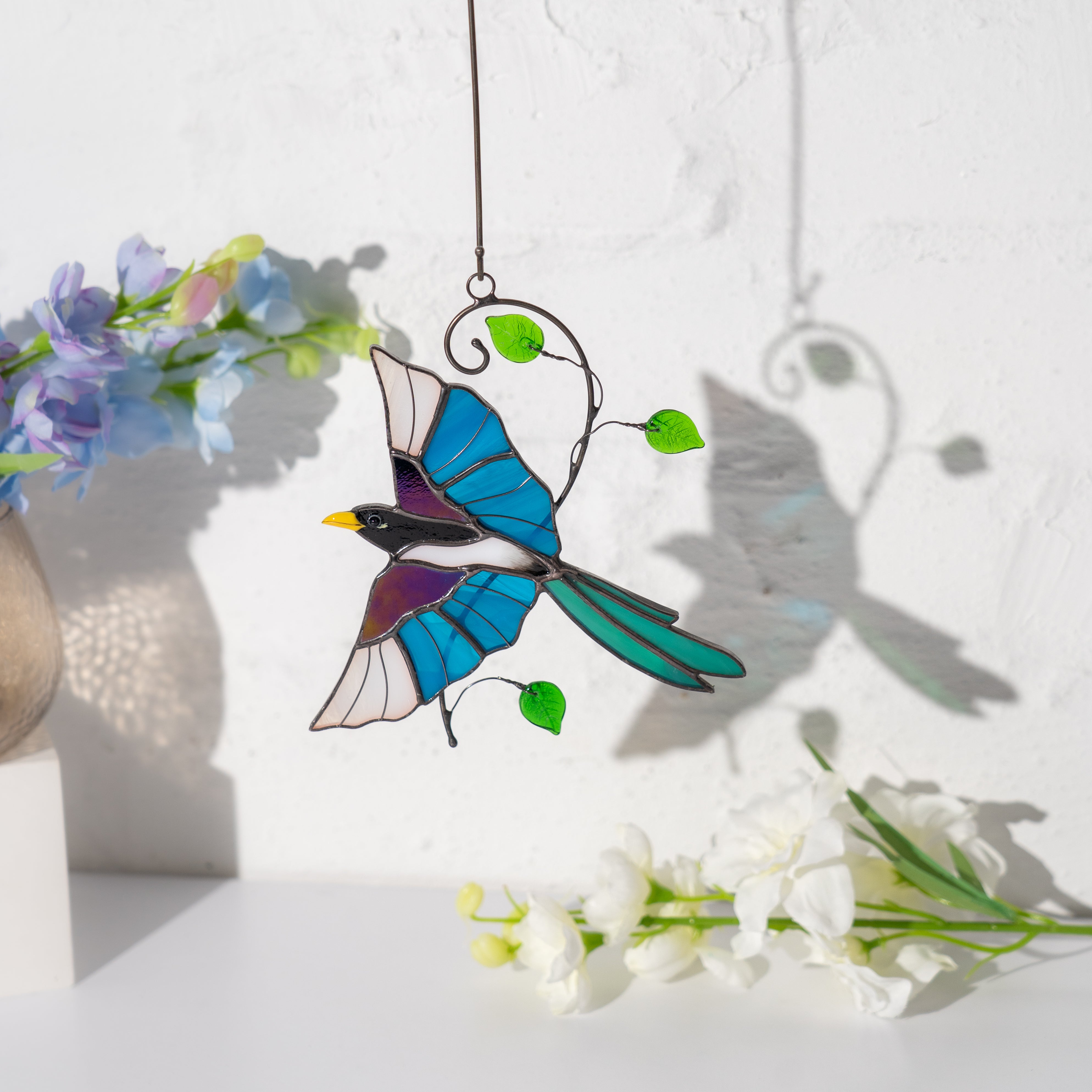 Stained glass magpie hanging by a white wall, casting a soft shadow, with pastel flowers nearby.