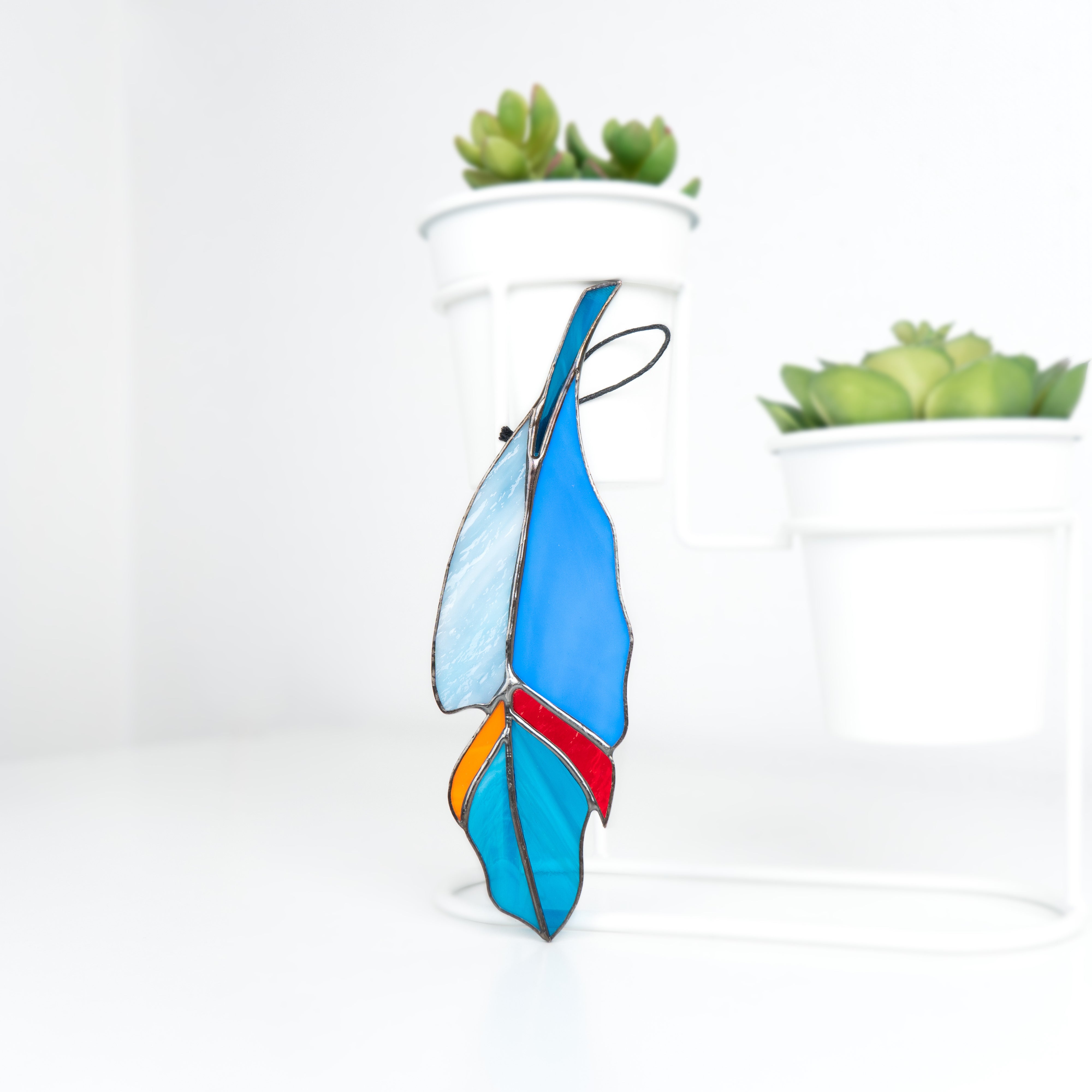 Blue-toned stained glass feather suncatcher standing upright on a white surface near potted succulents.