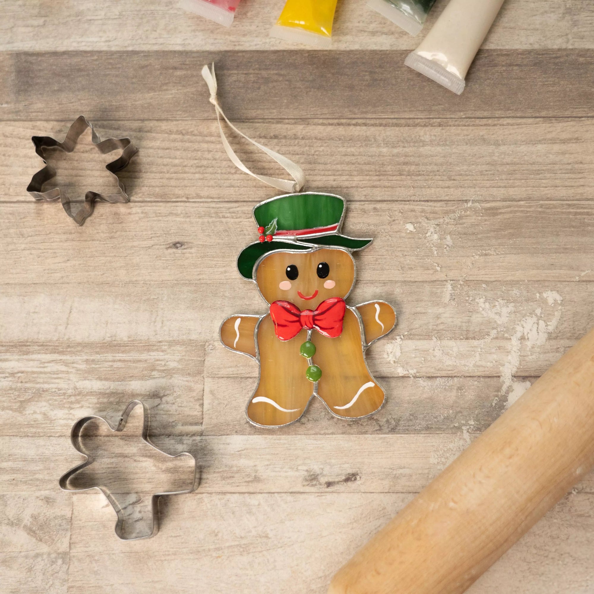Stained glass gingerbread man ornament placed beside cookie cutters and rolling pin on a wooden surface.