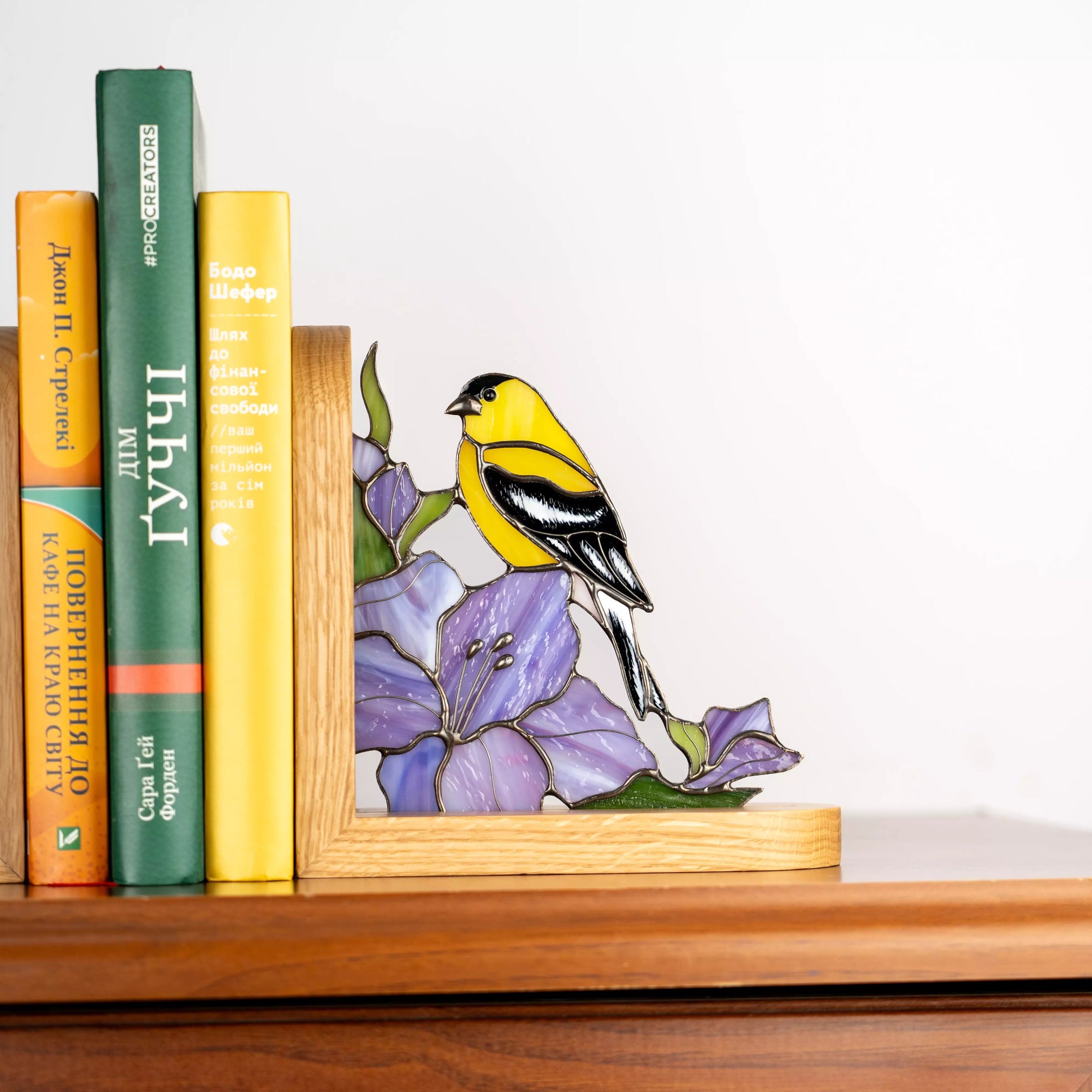 Stained glass goldfinch perched on a purple flower, handmade glass bookend displayed on a wooden shelf near books, combining functional book support with colorful bird-themed decor