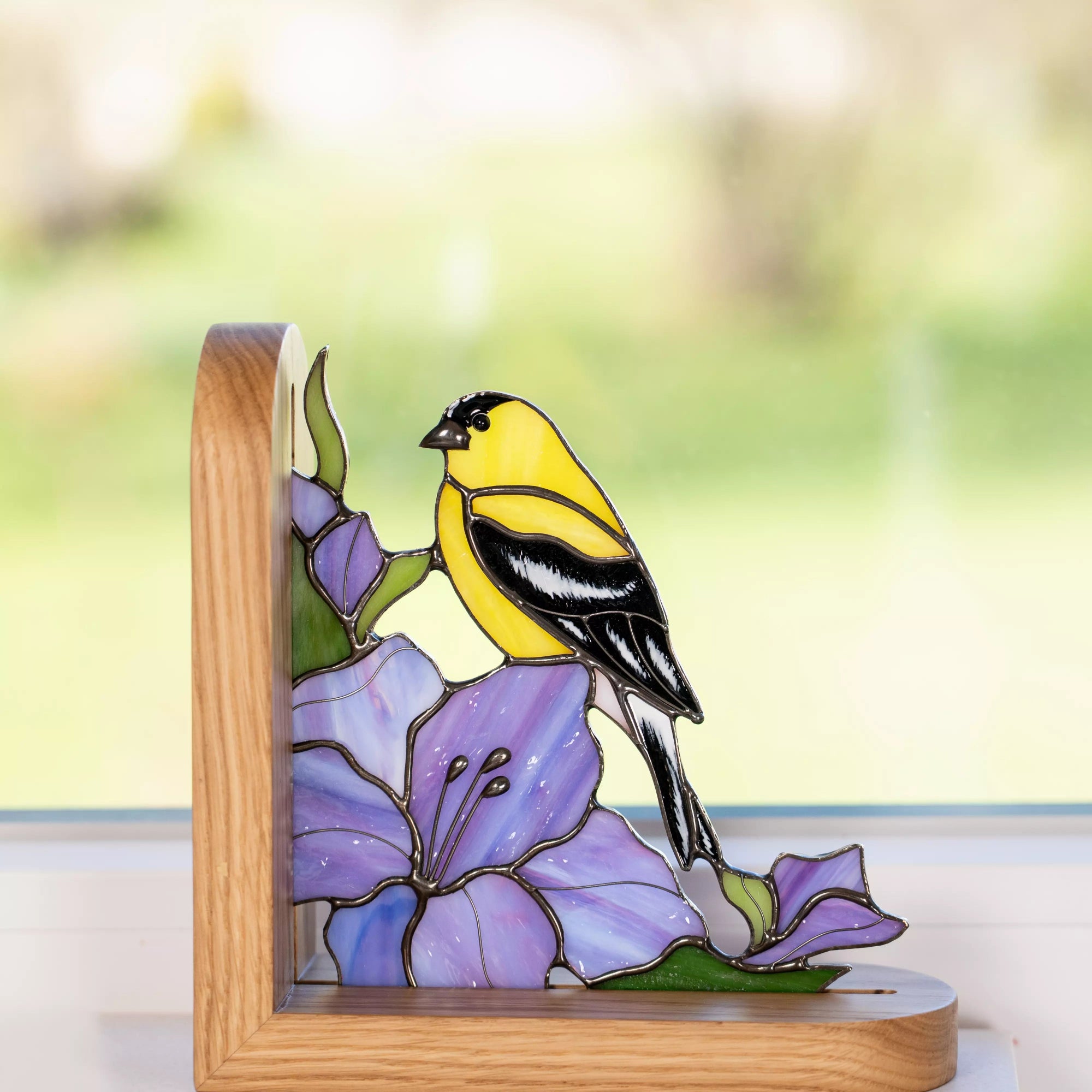 Stained glass panel with a yellow and black goldfinch on a purple flower, set against a blurred natural background.