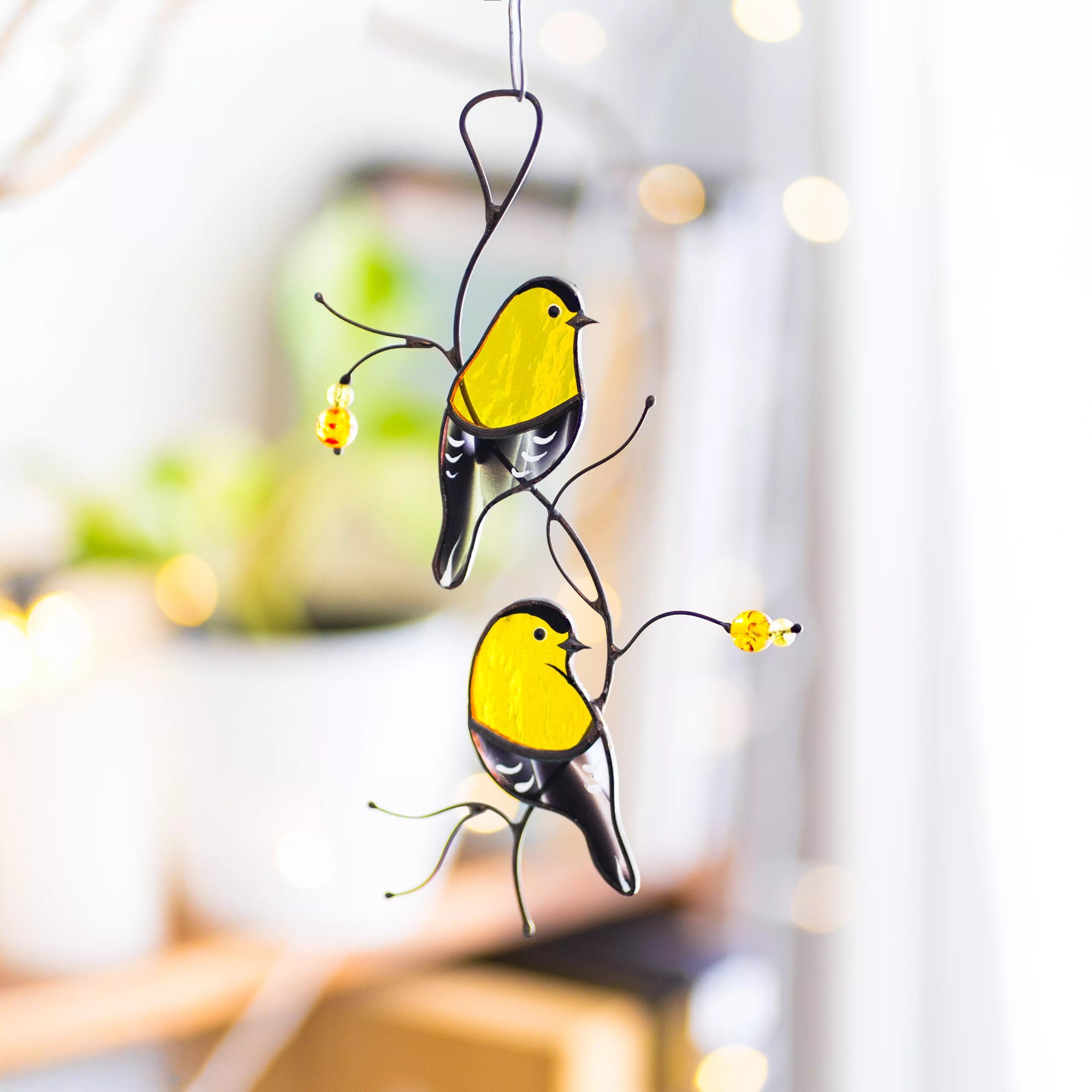A beautiful lifestyle shot of the goldfinch suncatcher hanging indoors with soft bokeh lighting and indoor plants in the background.