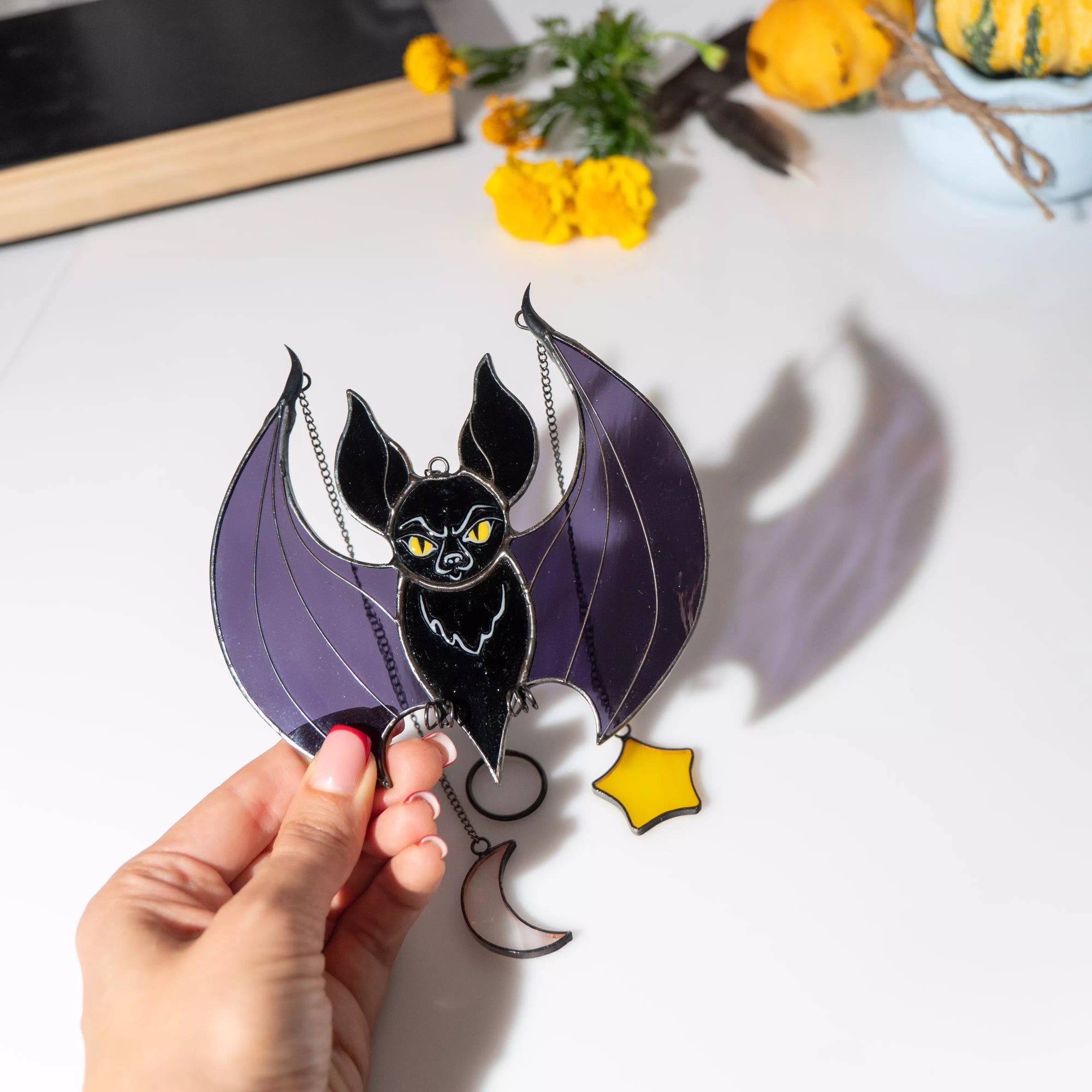 Hand holding a decorative bat with purple wings and yellow eyes on a white surface with flowers in the background.