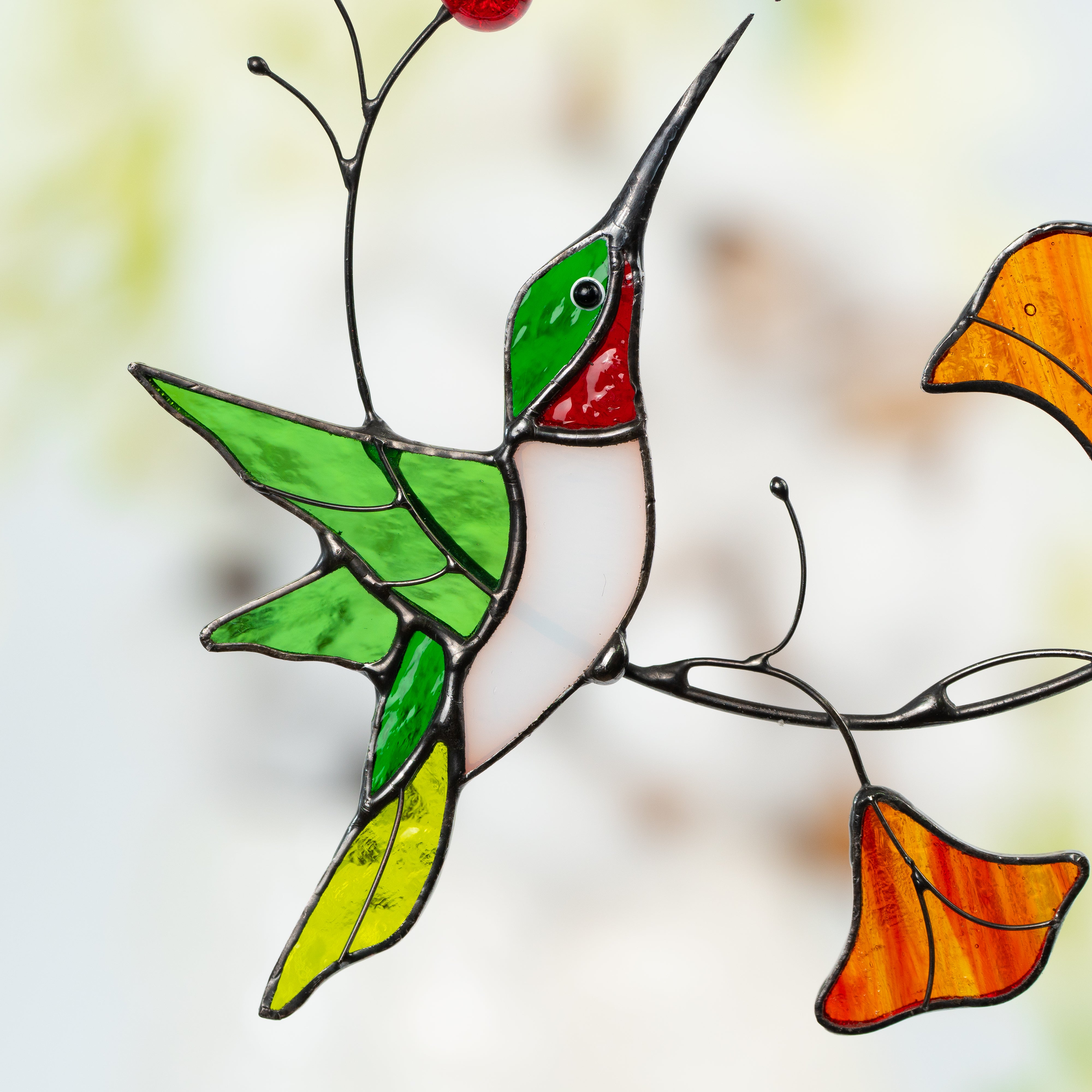 Close-up of stained glass hummingbird in green, red, and white flying toward an orange flower