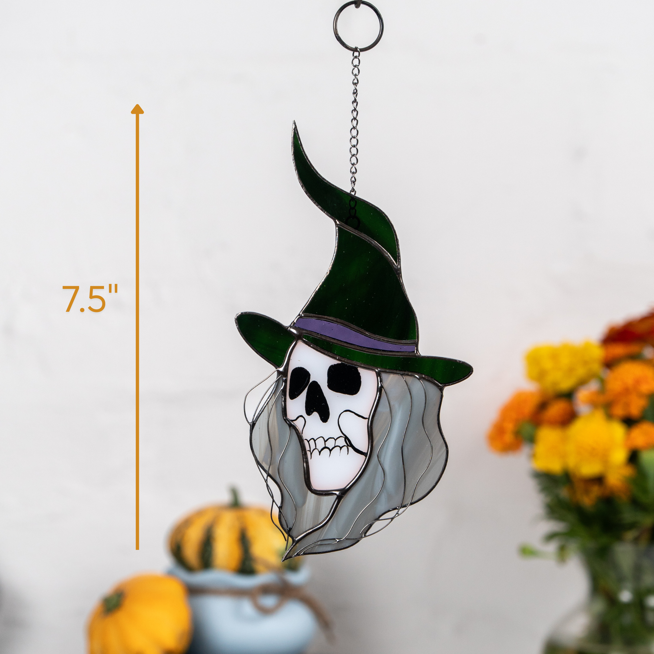 Witch skull suncatcher sized 7.5 inches with green pointy hat, hanging from chain near Halloween props and pumpkins