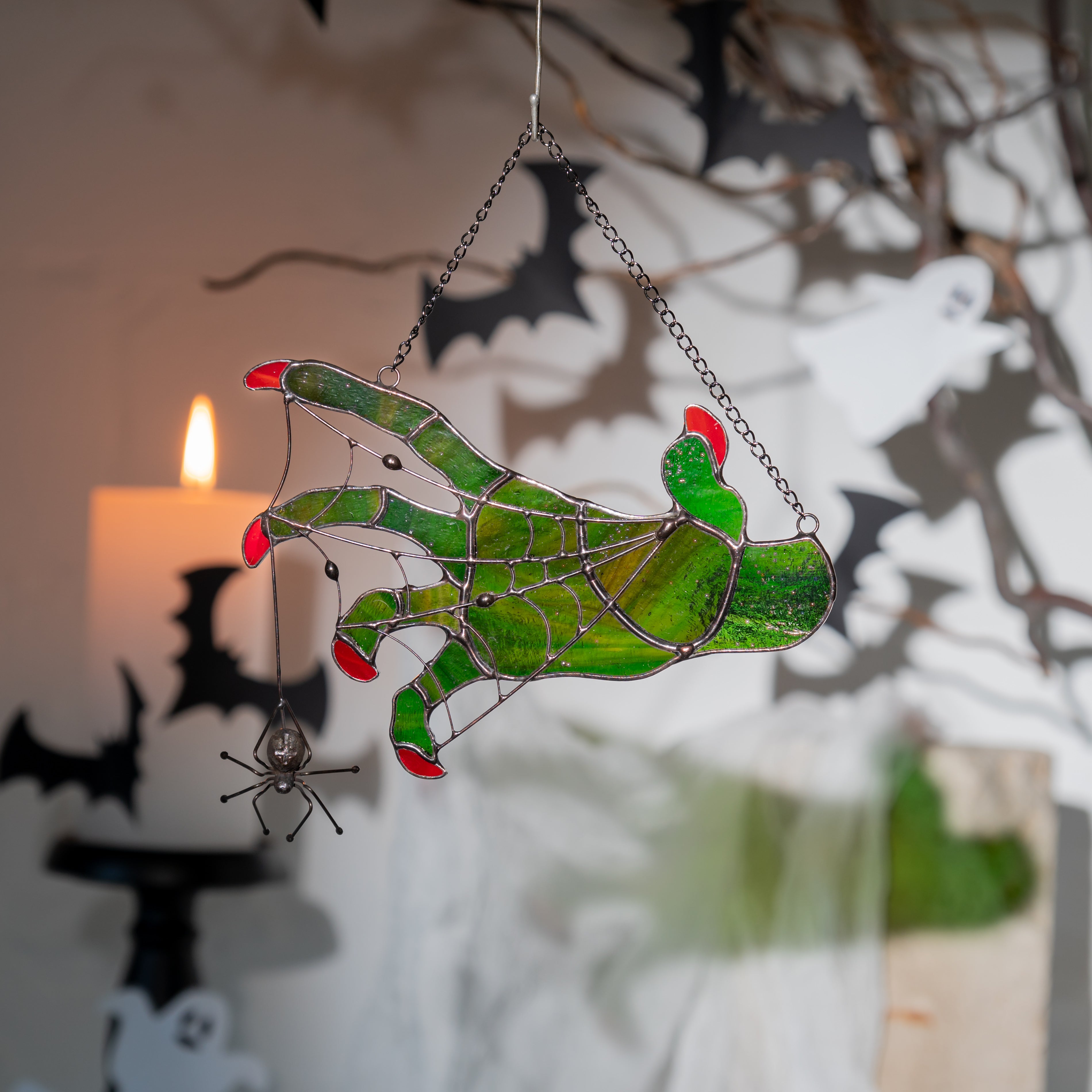 Witch hand suncatcher hanging indoors with Halloween props like bats, candle, and ghost silhouettes.