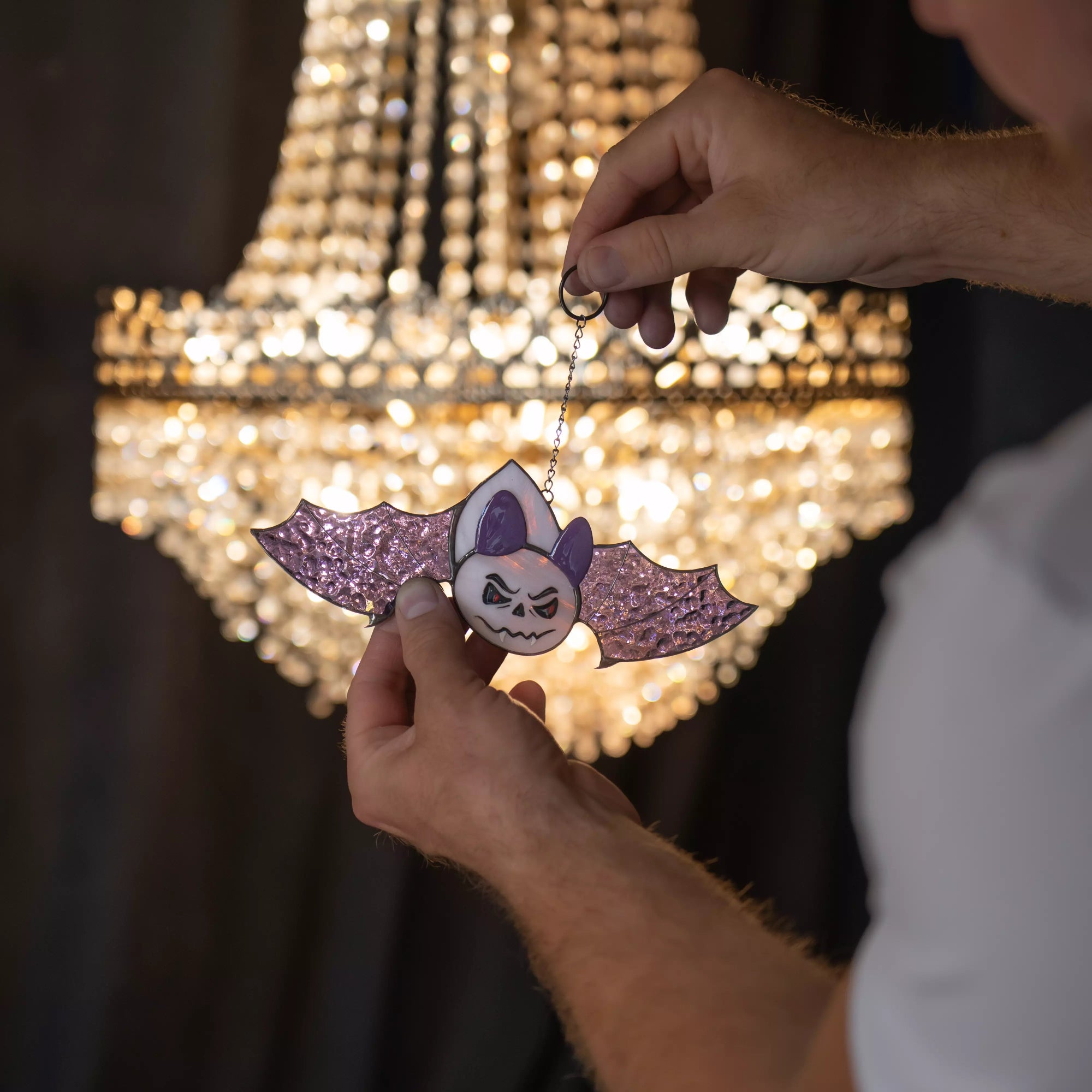 Person holding stained glass halloween bat suncatcher near chandelier light reflections