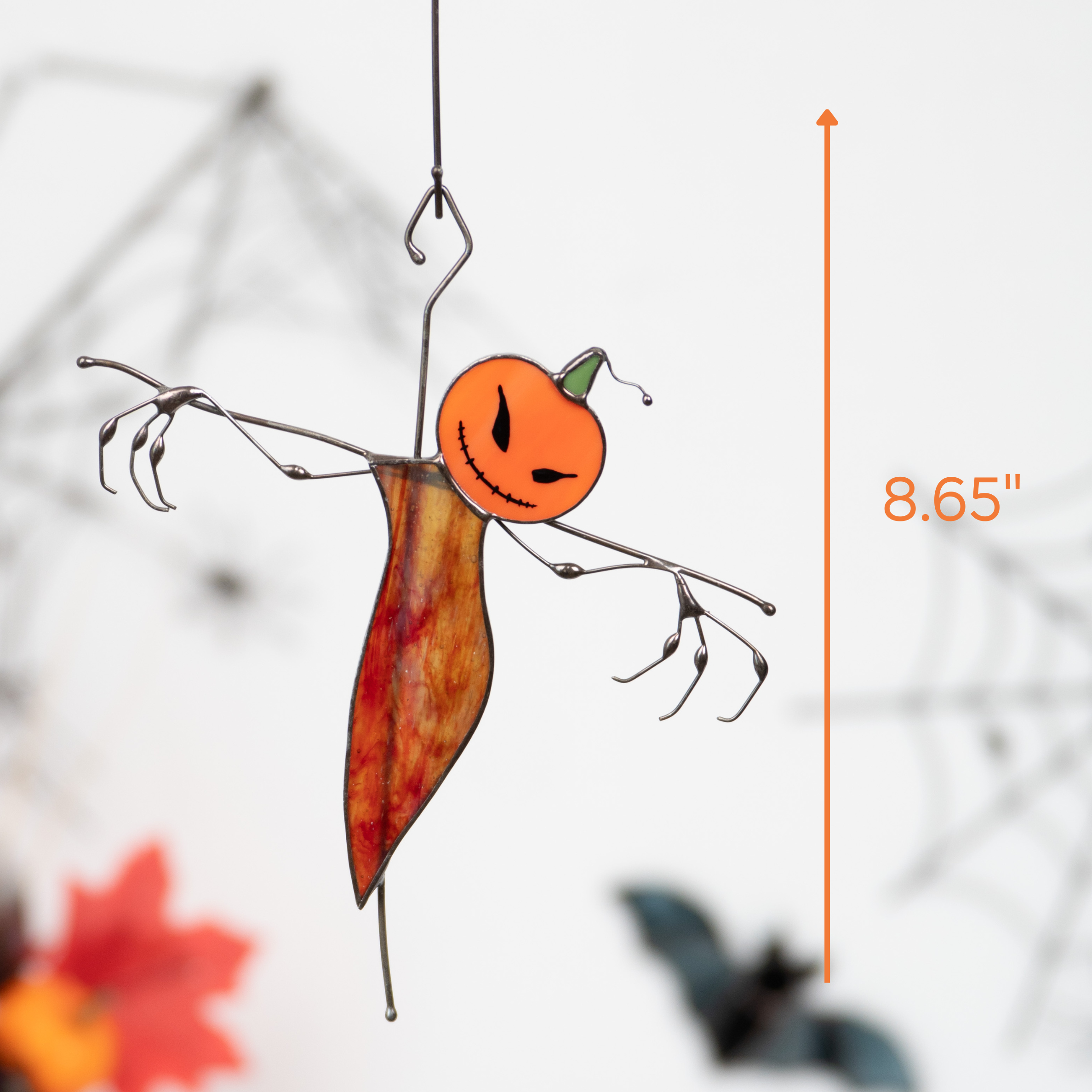 Stained glass scarecrow with a pumpkin head and wire arms hanging near spiderweb decorations.
