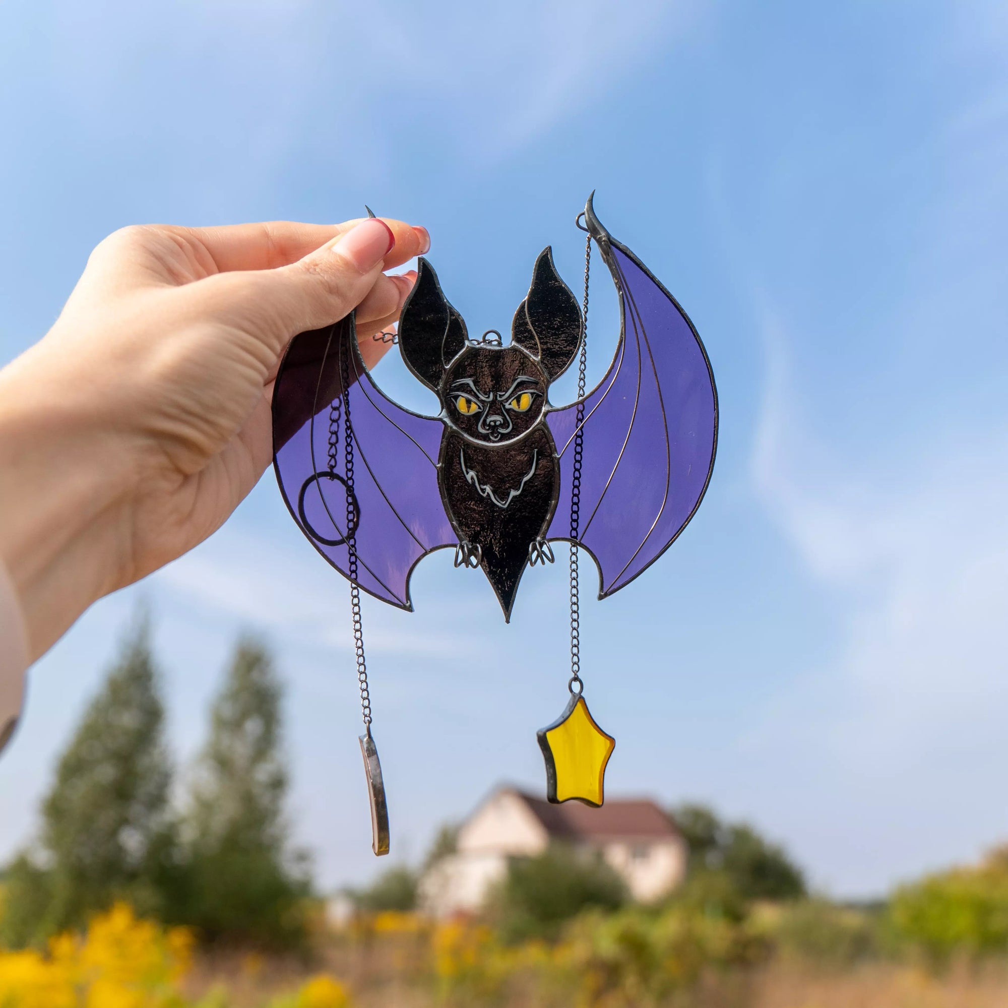 Hand holding black stained glass bat with moon and star charms against blue sky
