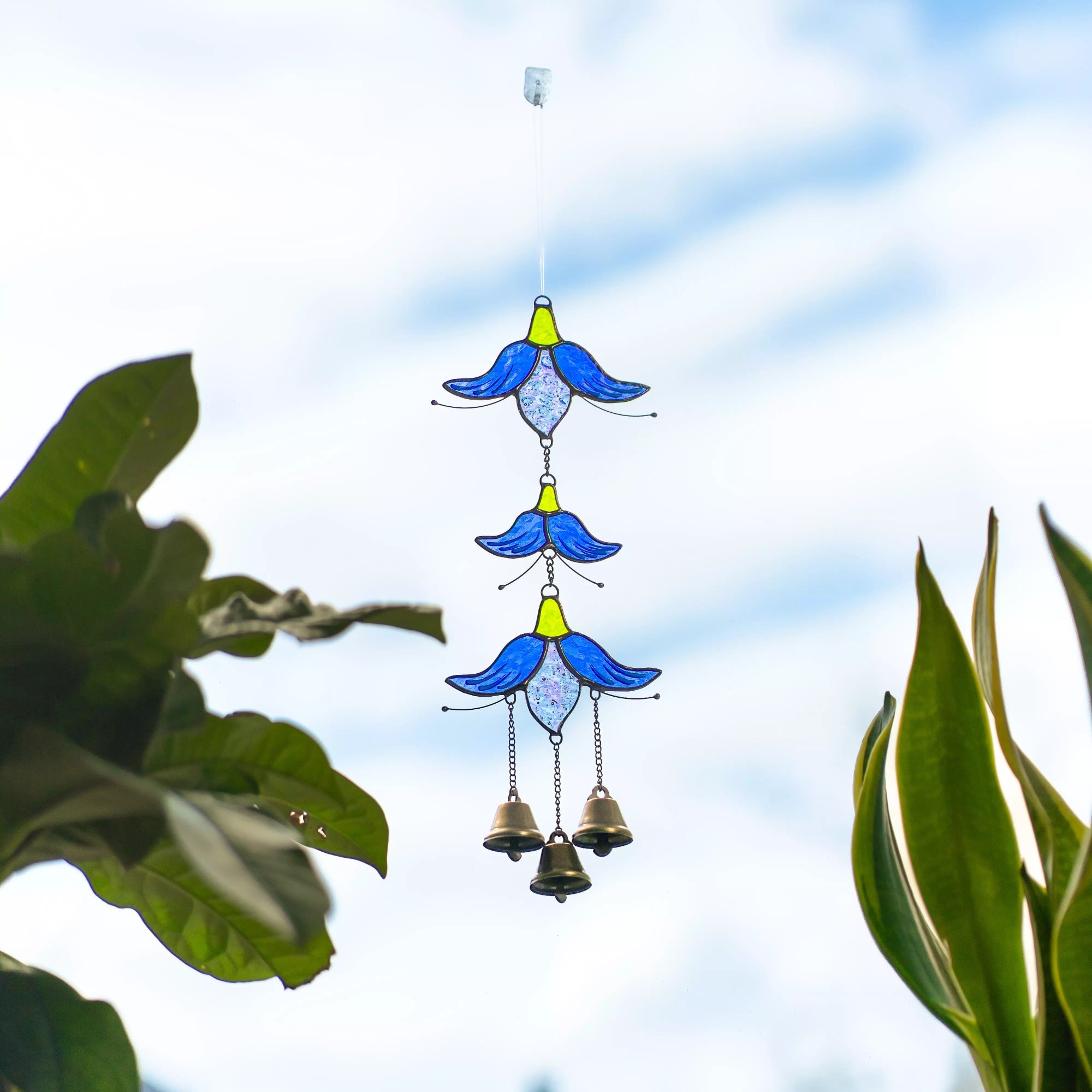 A person’s hand holding a blue and green stained glass flower wind chime in front of a window with indoor plants.