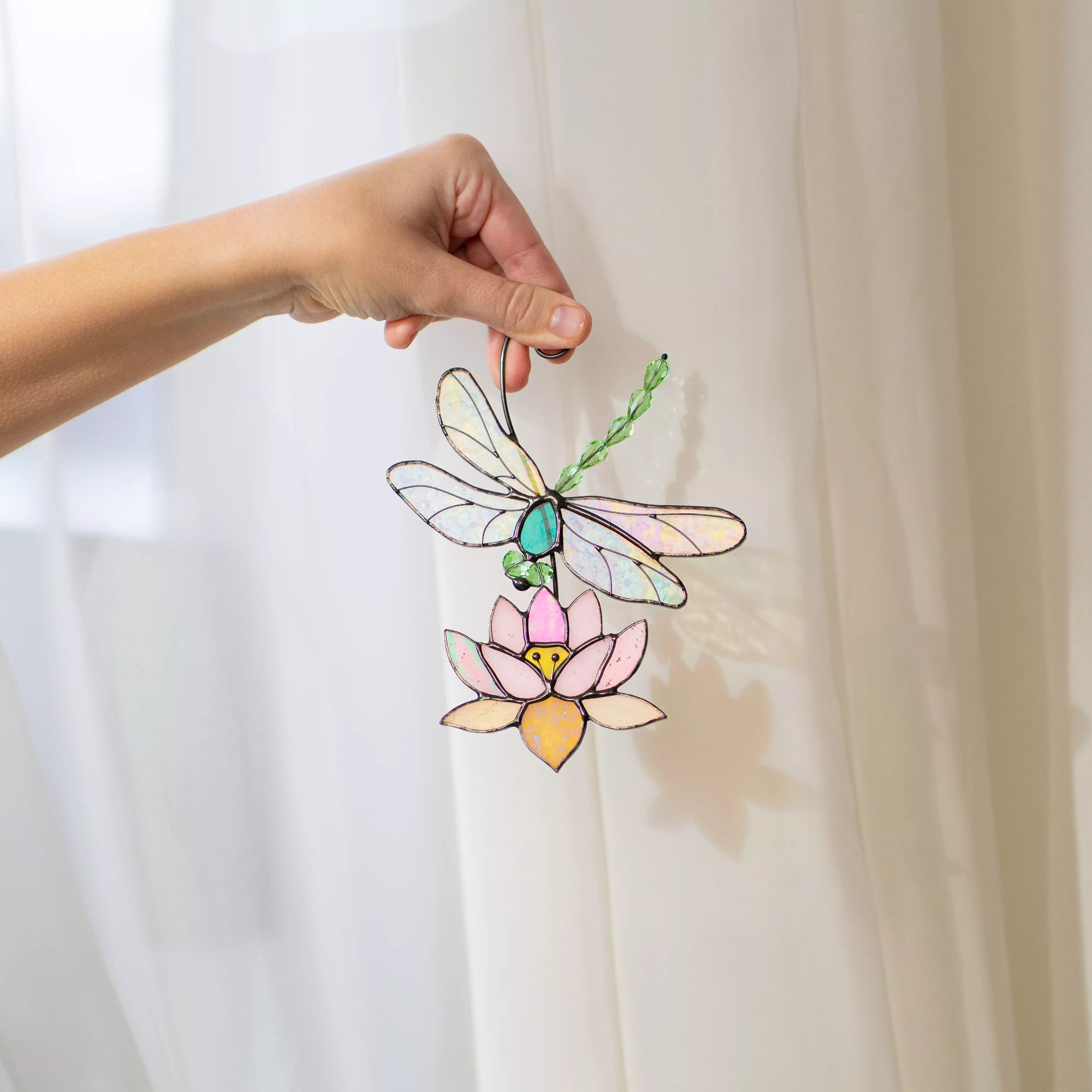 Hand holding a stained glass suncatcher dragonfly and lotus flower suncatcher against a white curtain.
