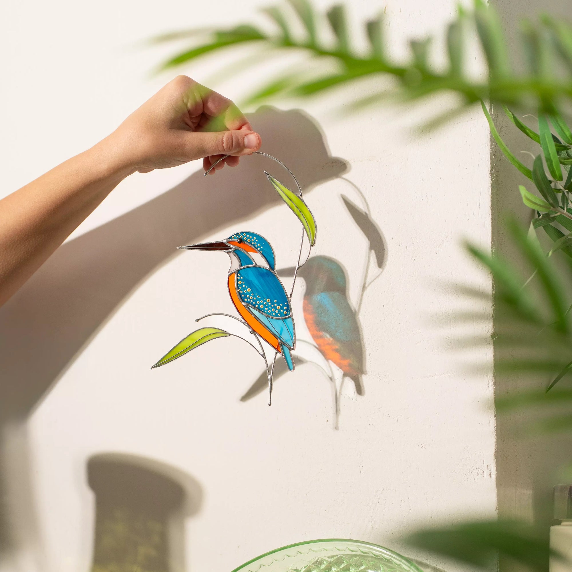 Person holding colorful stained glass kingfisher bird decoration with green leaves against a light background.