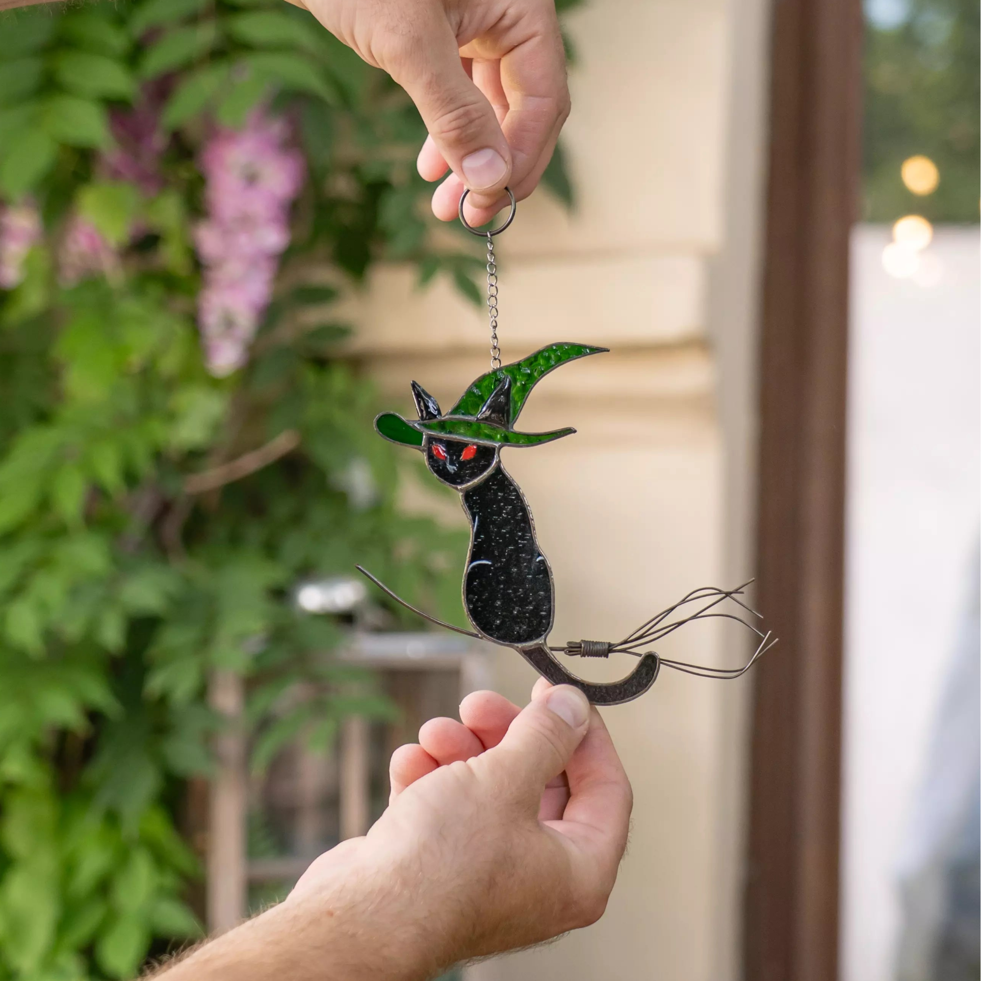 Hand holding a stained glass black cat ornament with green leaves against a blurred outdoor background.