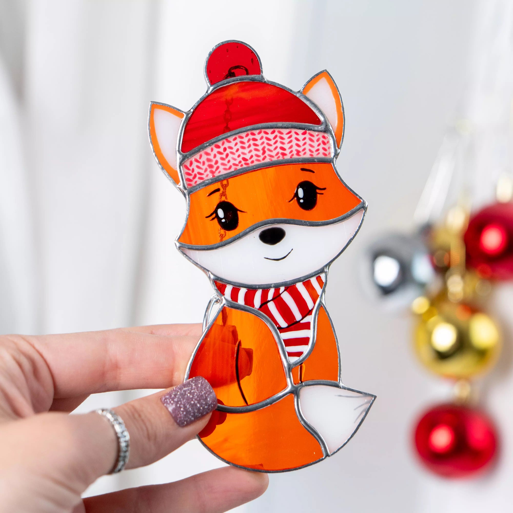 Hand holding a decorative stained glass fox ornament with a red hat and scarf against a blurred background of Christmas decorations.