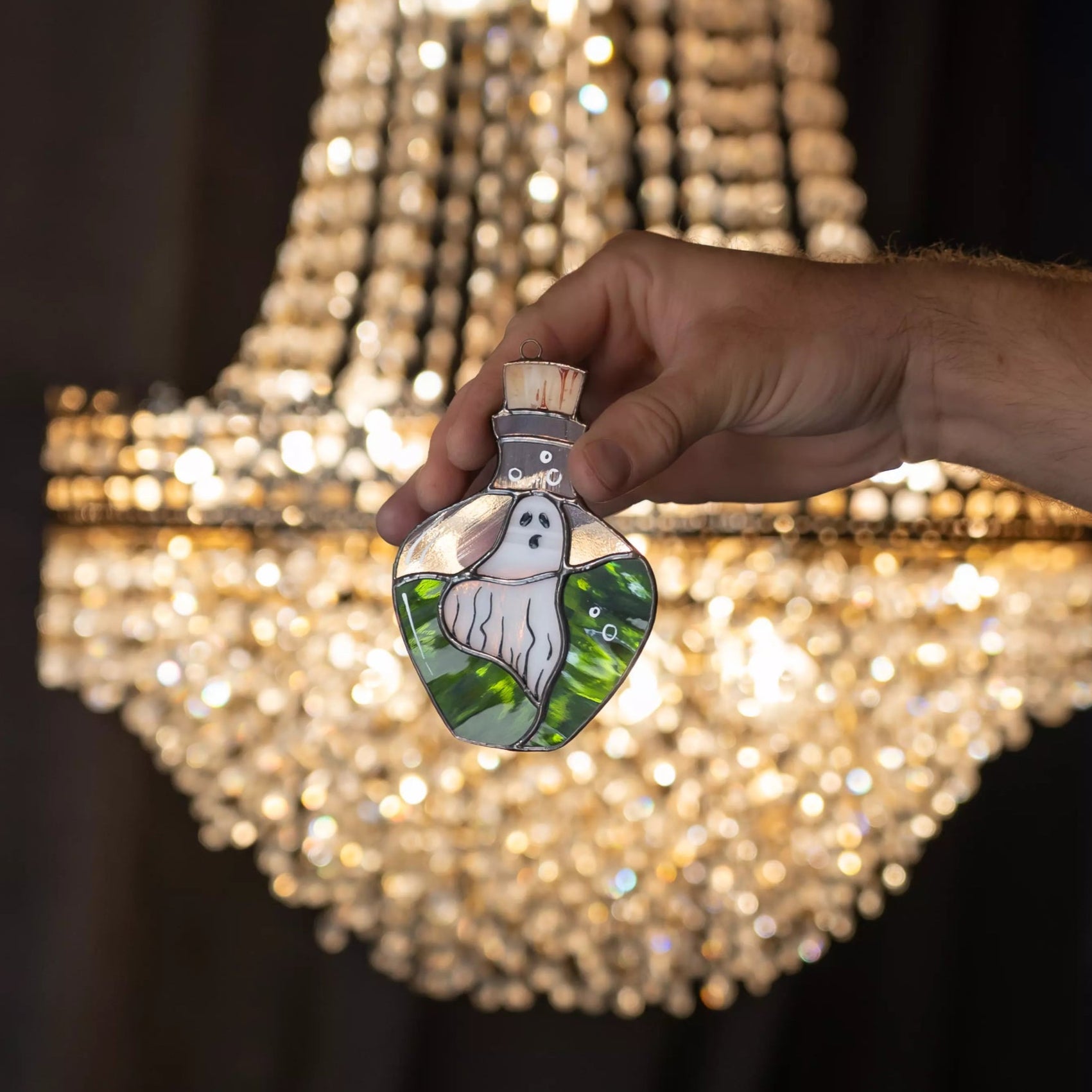 Hand holding a decorative stained glass ghost in bottle suncacther in front of a sparkling chandelier.