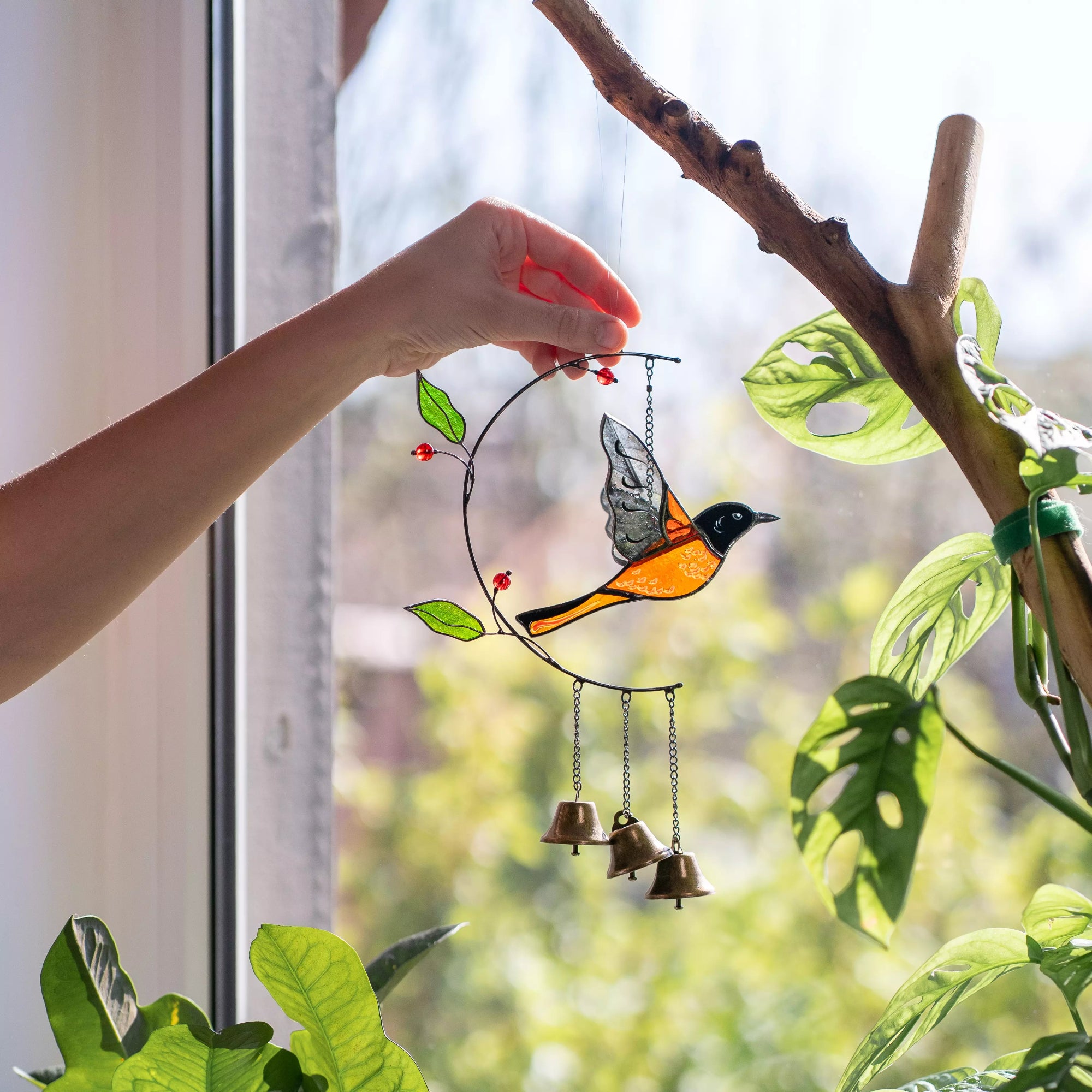 Hand holding a stained glass wind chime with a baltimore oriole design near a window with plants.
