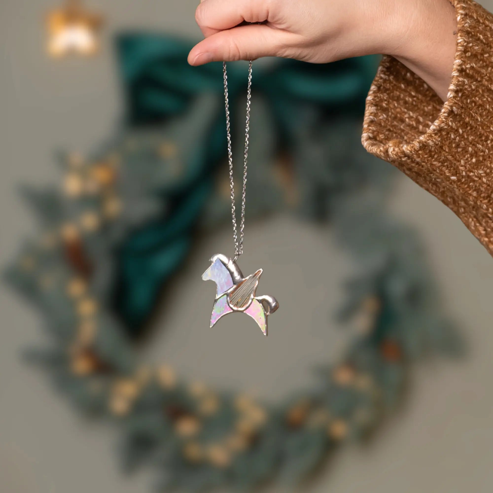 Person holding a necklace with an iridescent stained glass pegasus pendant in front of a blurred decorative wreath.