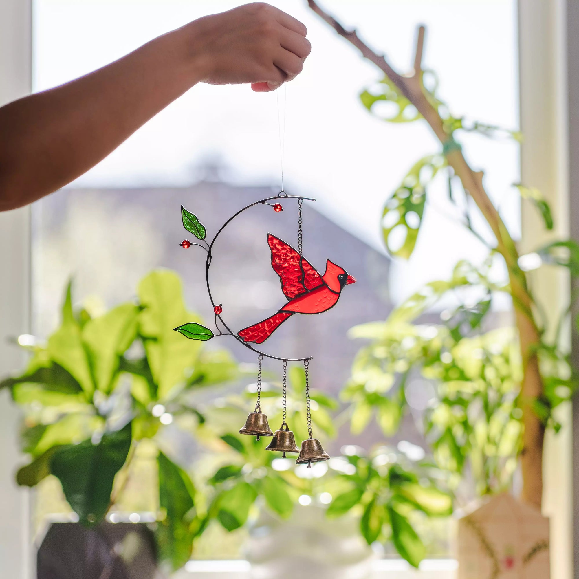 Hand holding a stained glass cardinal bird-shaped glass ornament with bells against a blurred indoor background