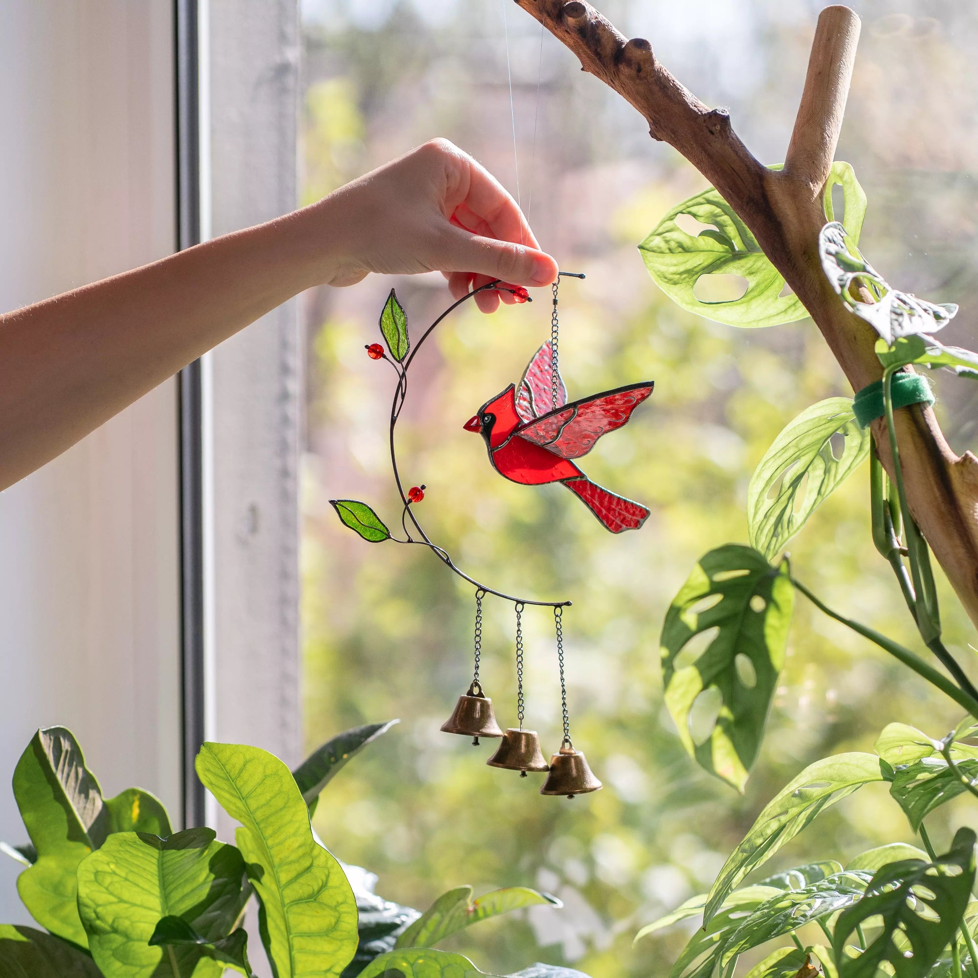 Hand holding stained glass cardinal bird ornament with bells near a window with greenery outside.