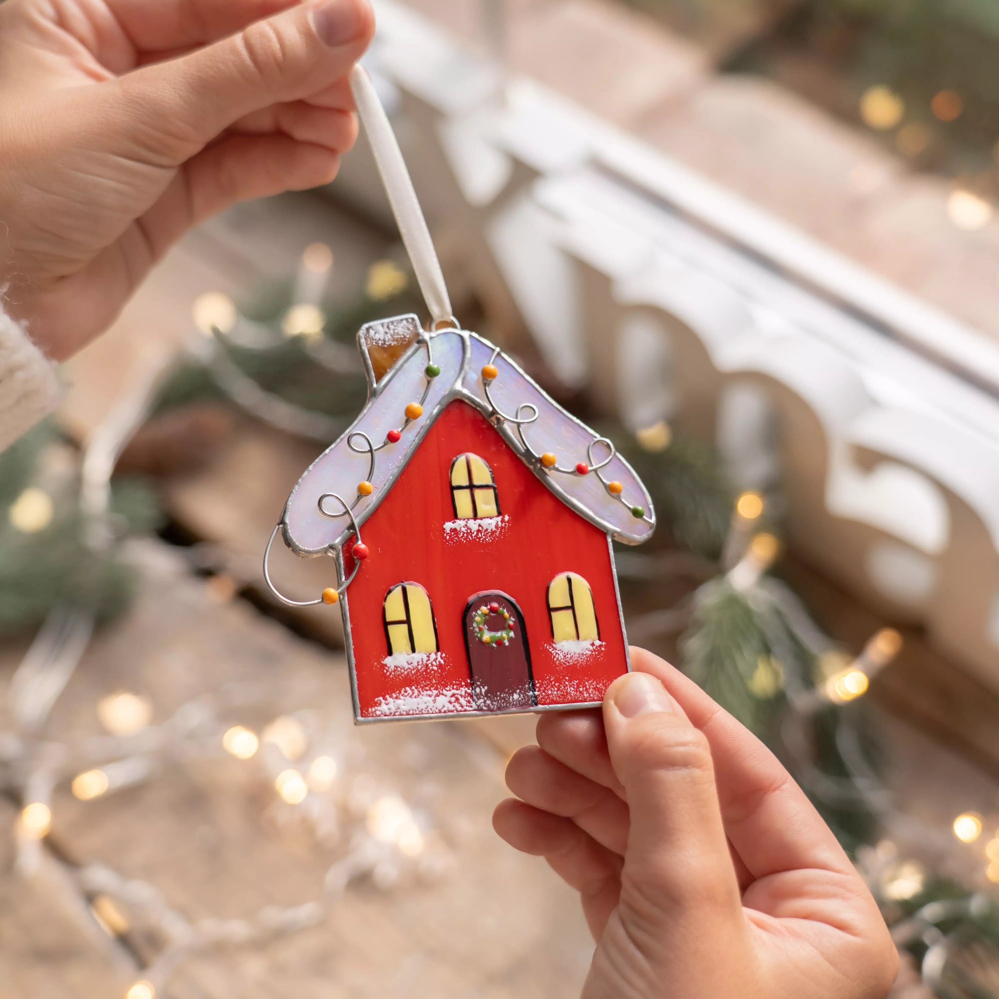 Hand holding a red house stained glass ornament with lights and decorations in the background