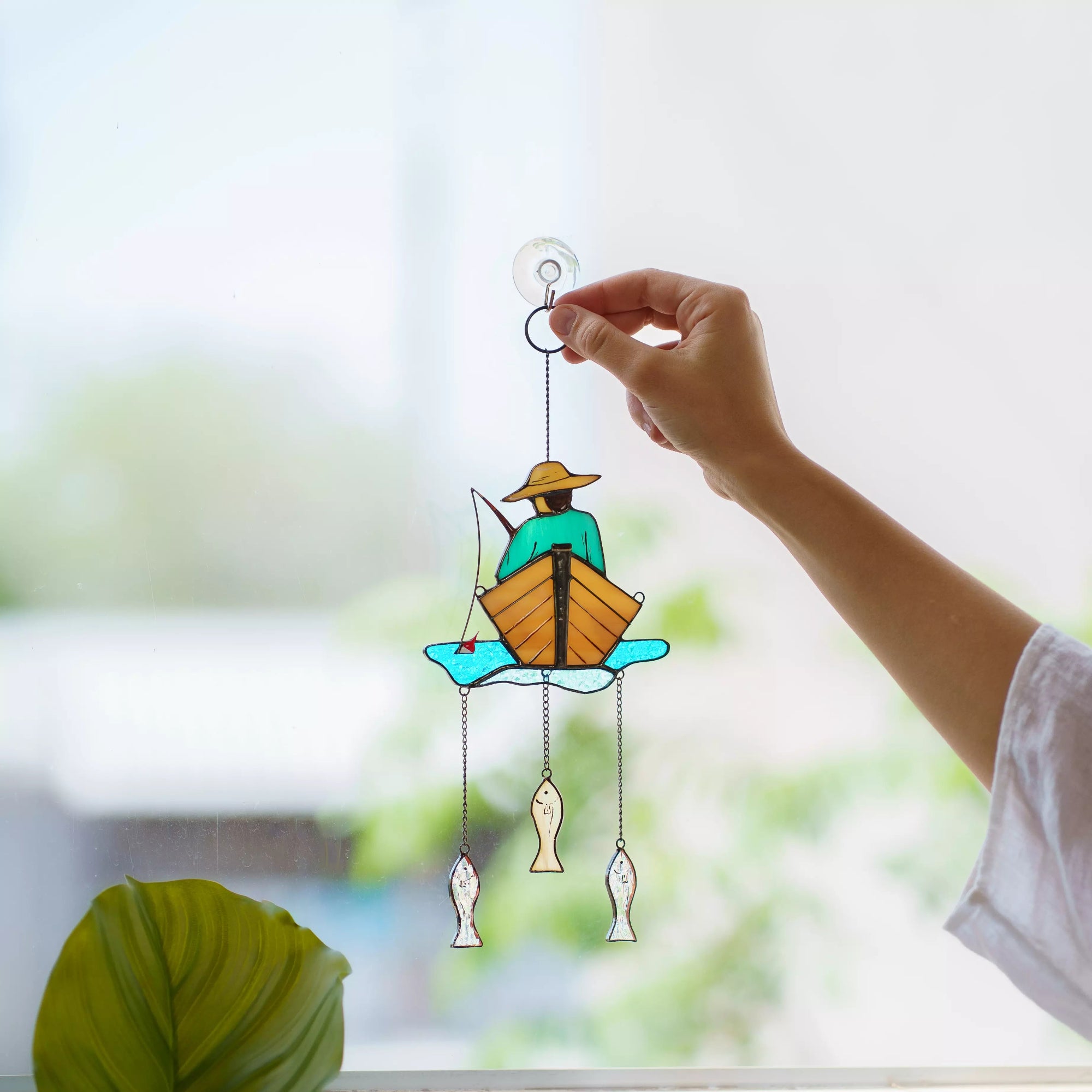 Hand holding a stained glass fisherman window hanging against a blurred window background
