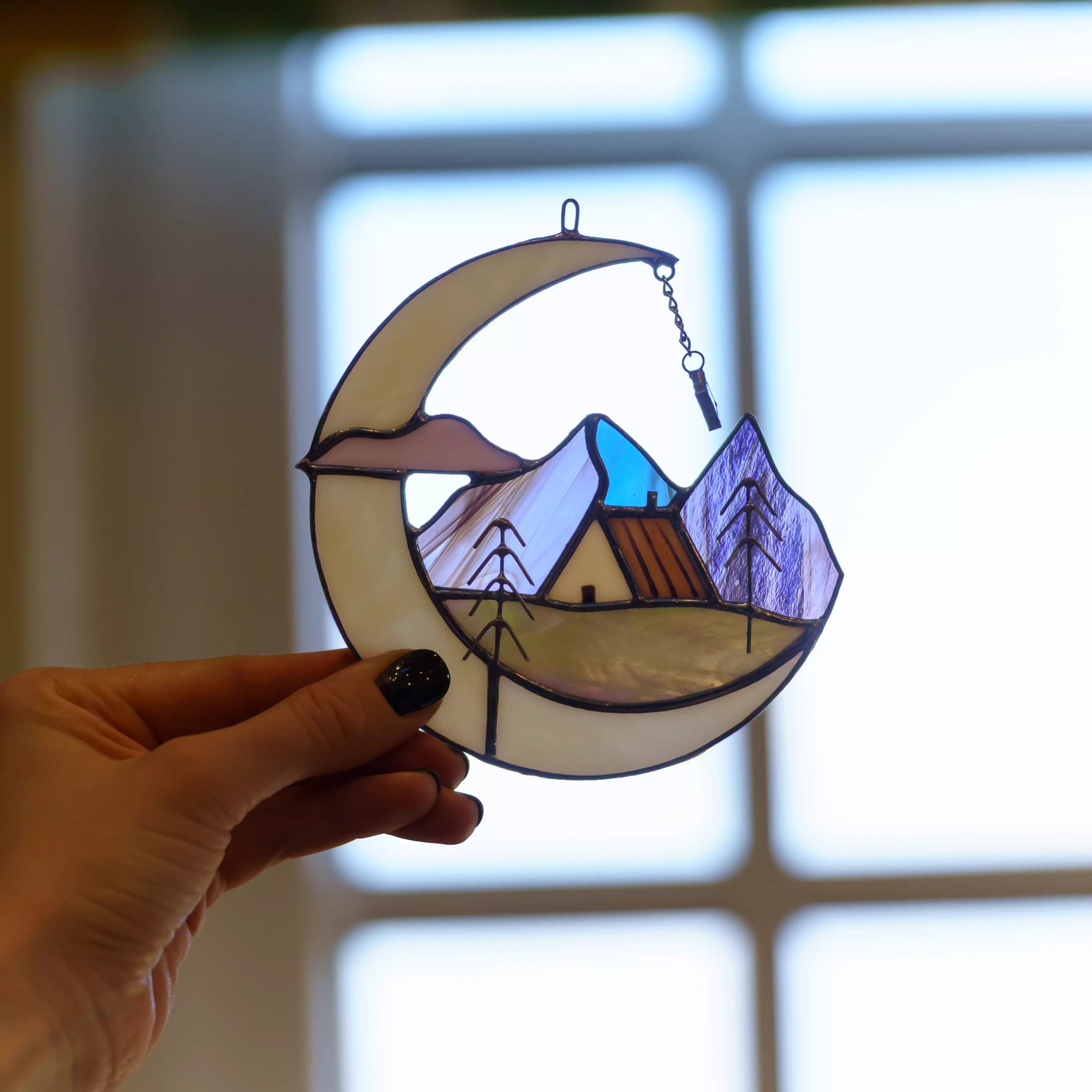 Hand holding a stained glass moon suncatcher with a house and trees design.