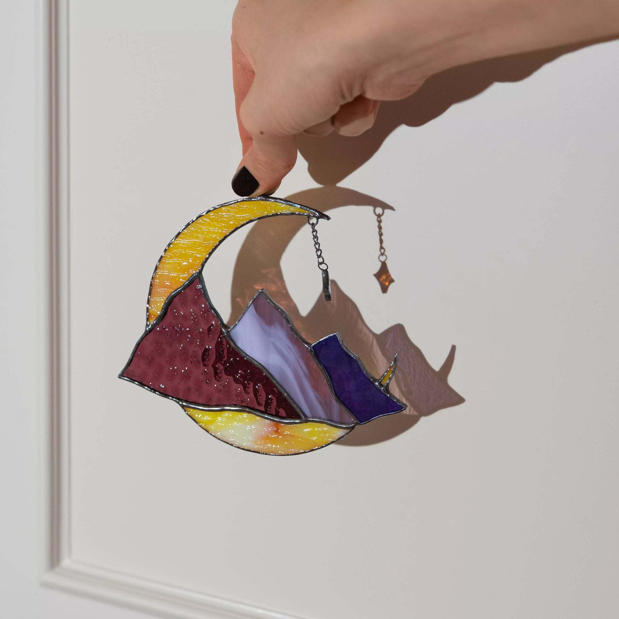 Hand holding a colorful stained glass suncatcher with mountains and a crescent moon.