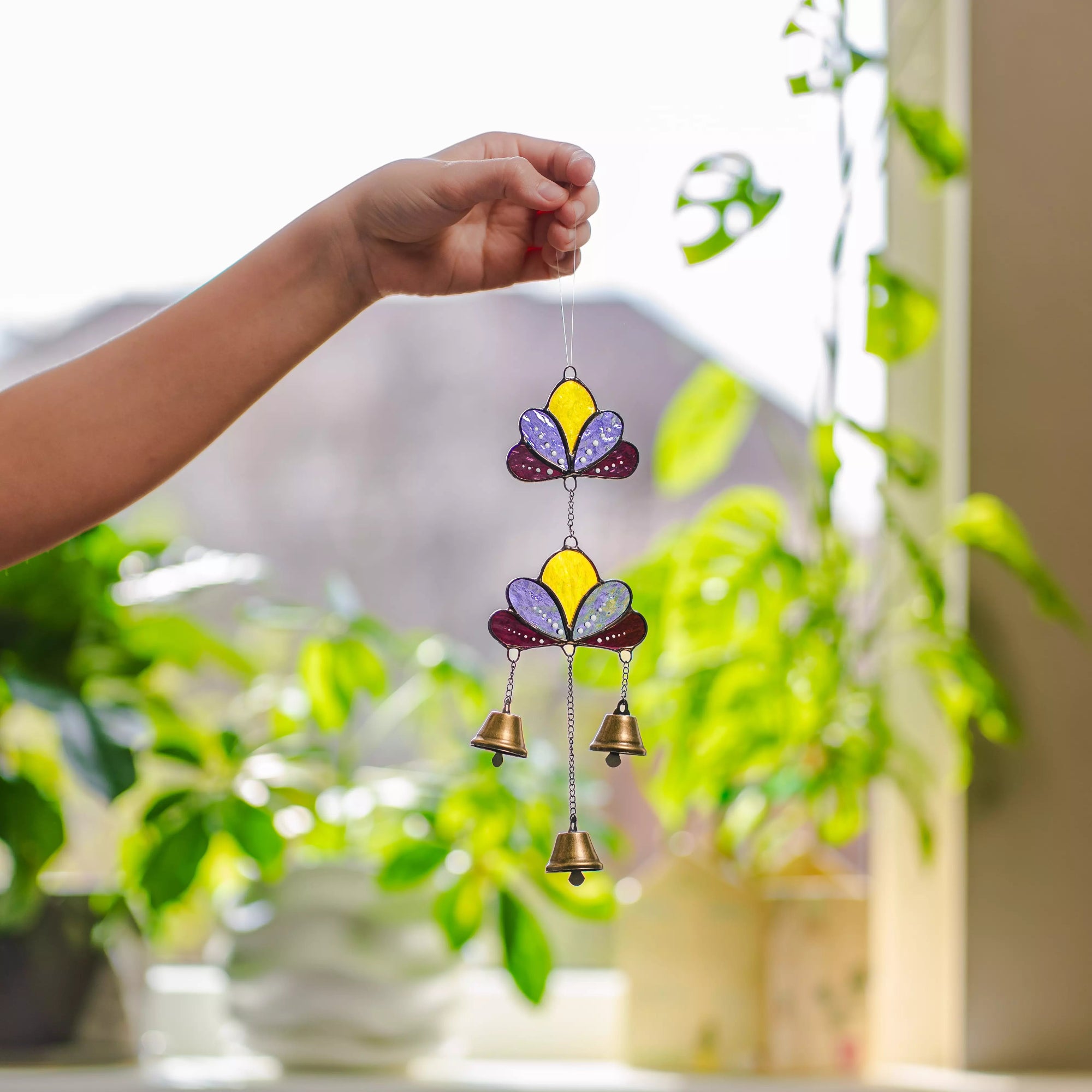 Hand holding a vibrant lotus-patterned stained glass wind chime against a bright, softly blurred green outdoor background.