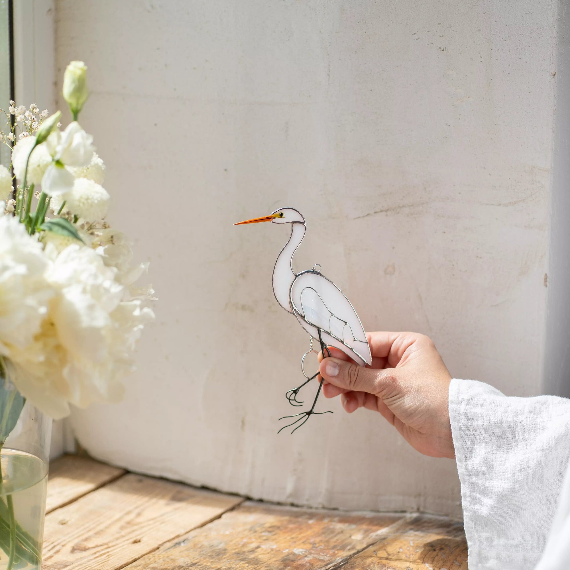 Hand holding stained glass heron next to a vase of white flowers on a wooden surface.