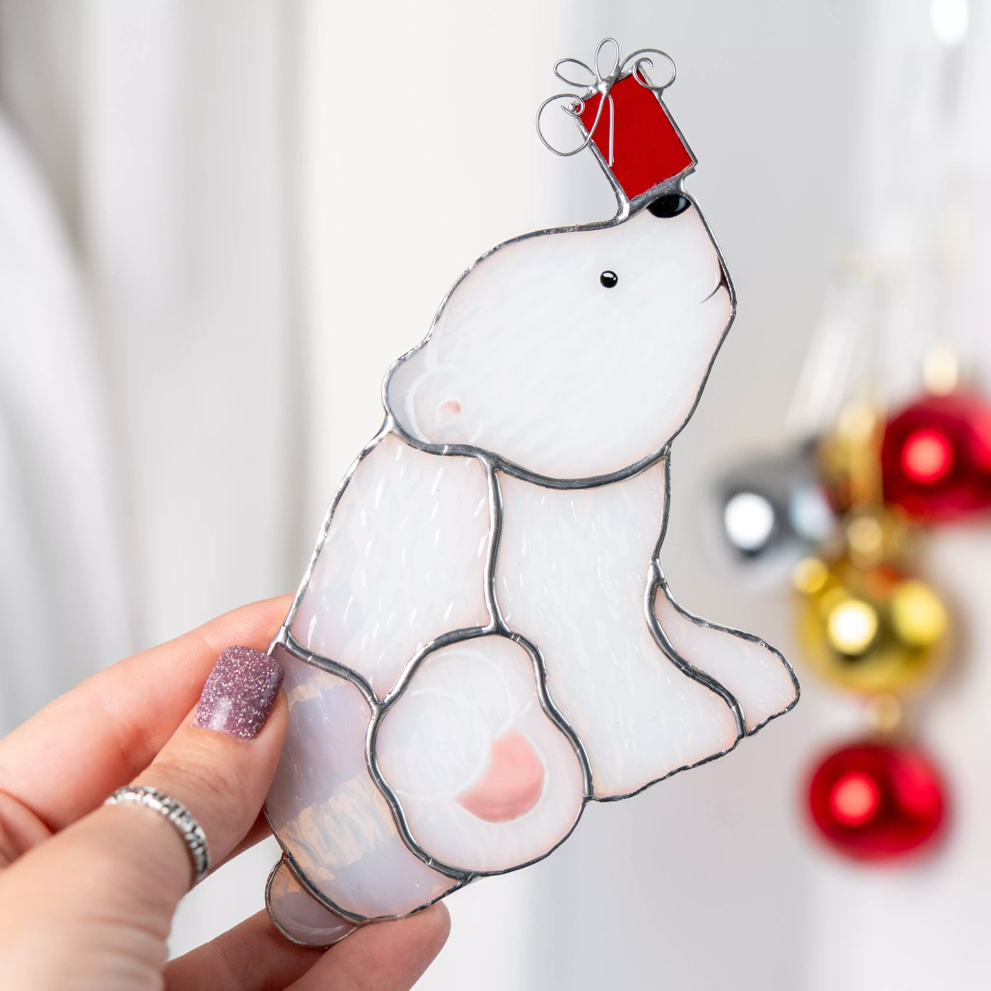Hand holding a white stained glass suncatcher featuring polar bear with a red gift on its head against a blurred background of Christmas decorations.