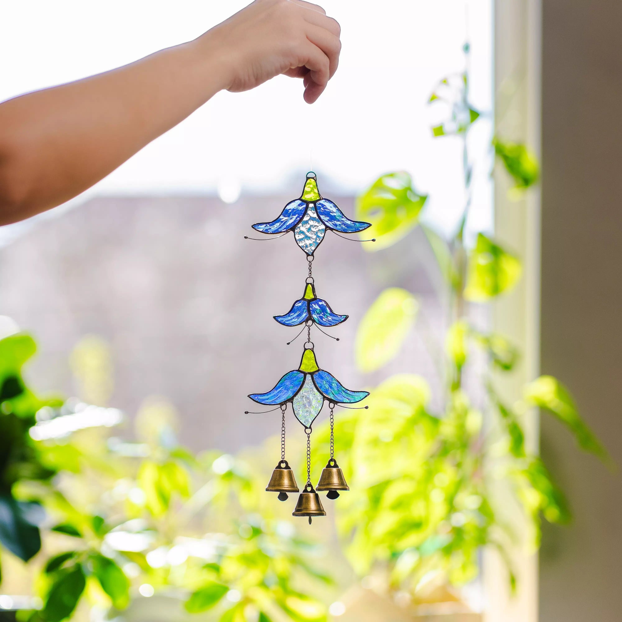 Hand holding a blue and green stained glass flower wind chime against a bright, softly blurred green outdoor background.