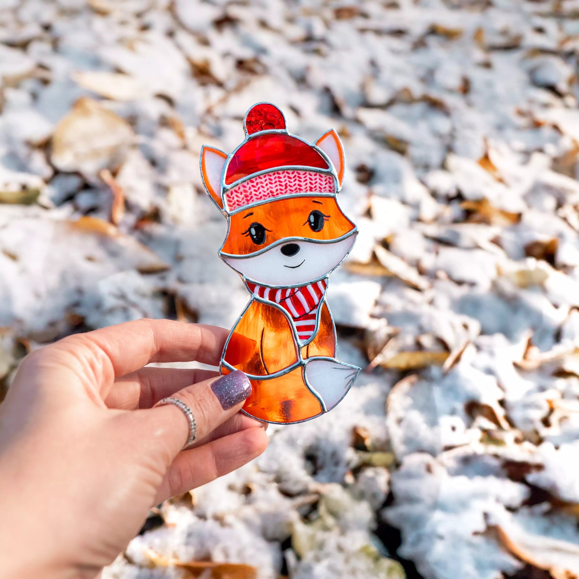 Hand holding stained glass suncatcher fox with a red hat and scarf against a snowy background