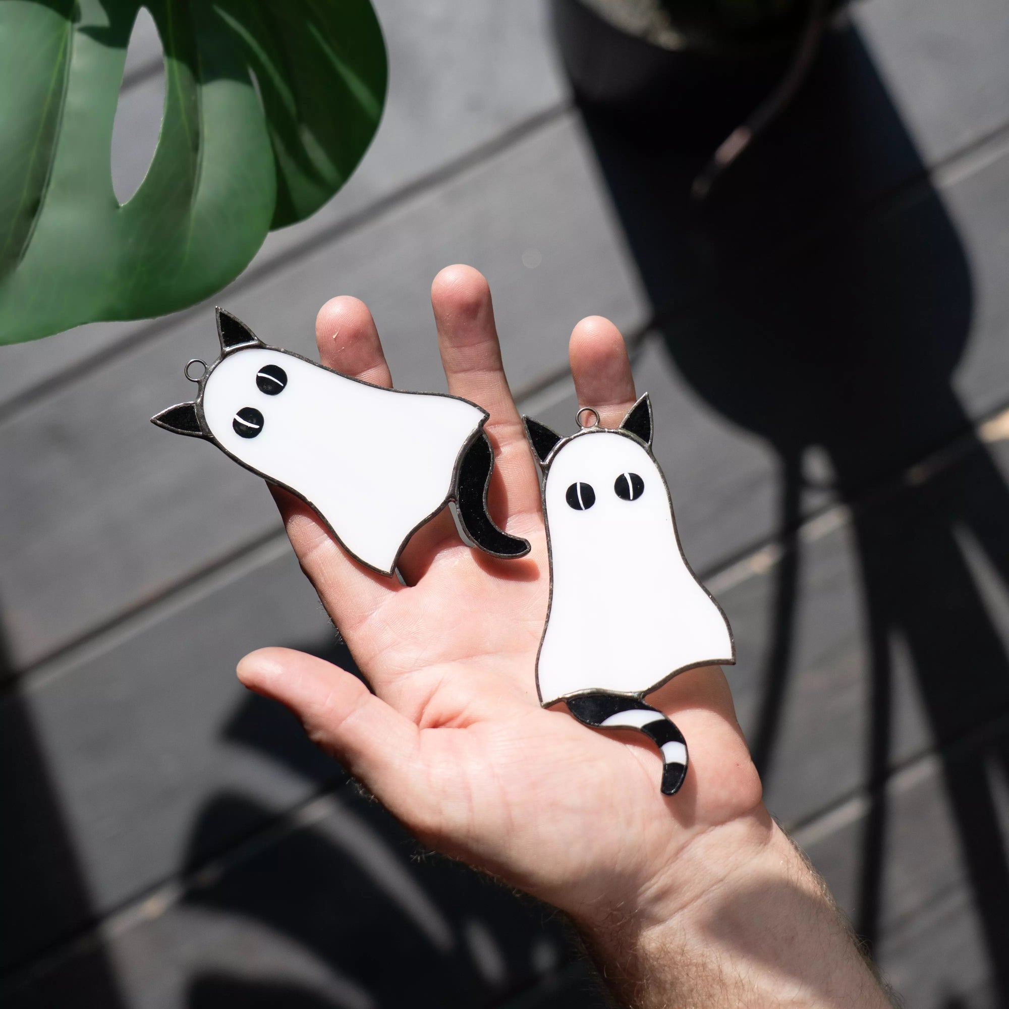Hand holding two black and white cat-shaped suncatchers against a dark surface with a plant leaf.