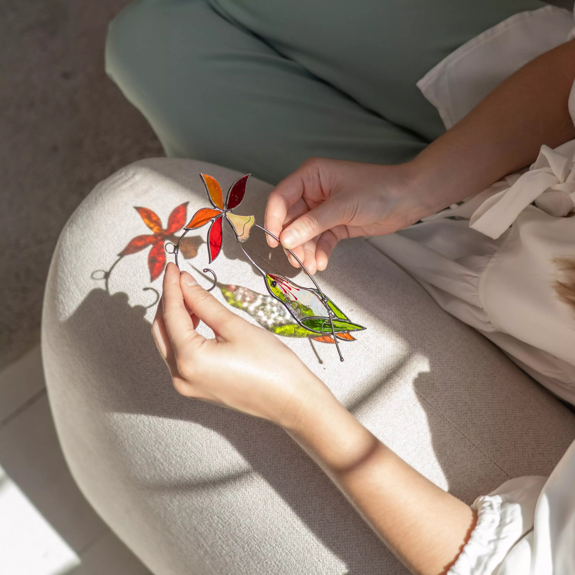 Person holds a colorful stained glass hummingbird with bell flower suncatcher, that casts a soft shadow