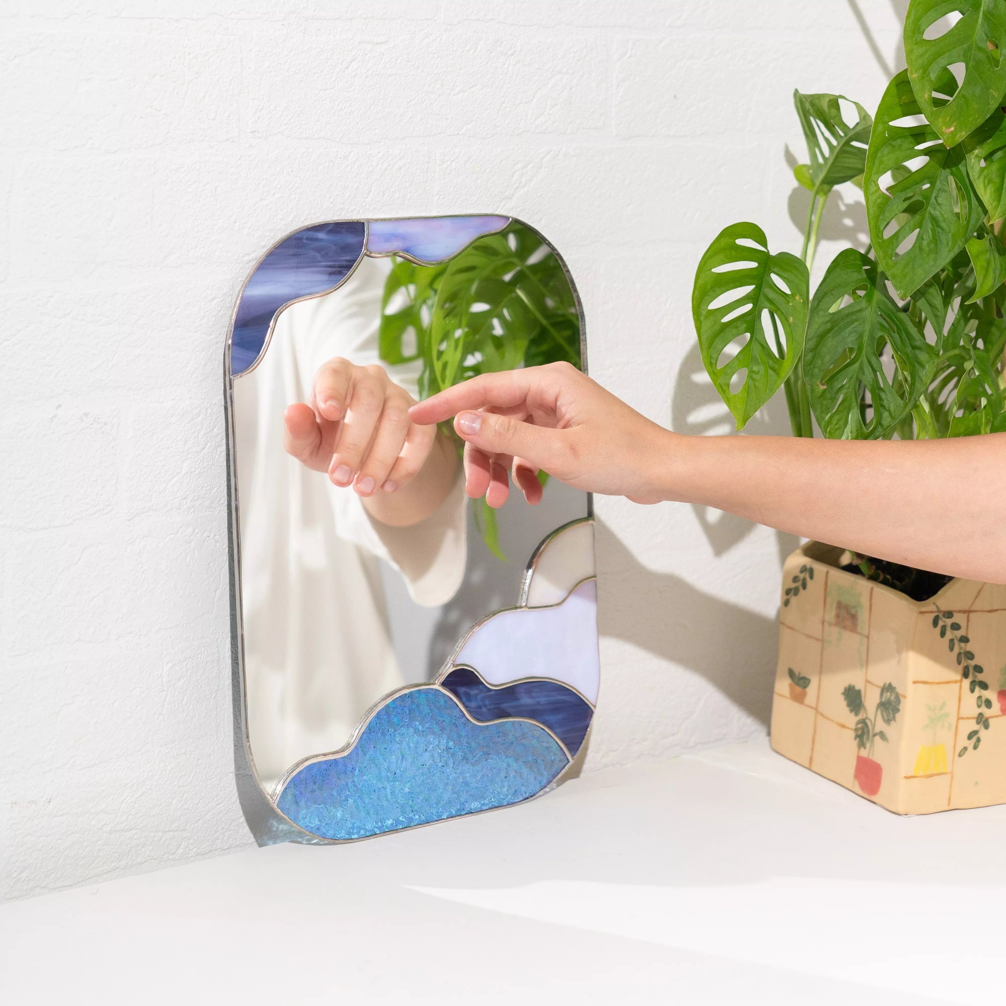 Hand reaching towards a stained glass mirror with cloud and mountain design, next to a potted plant.