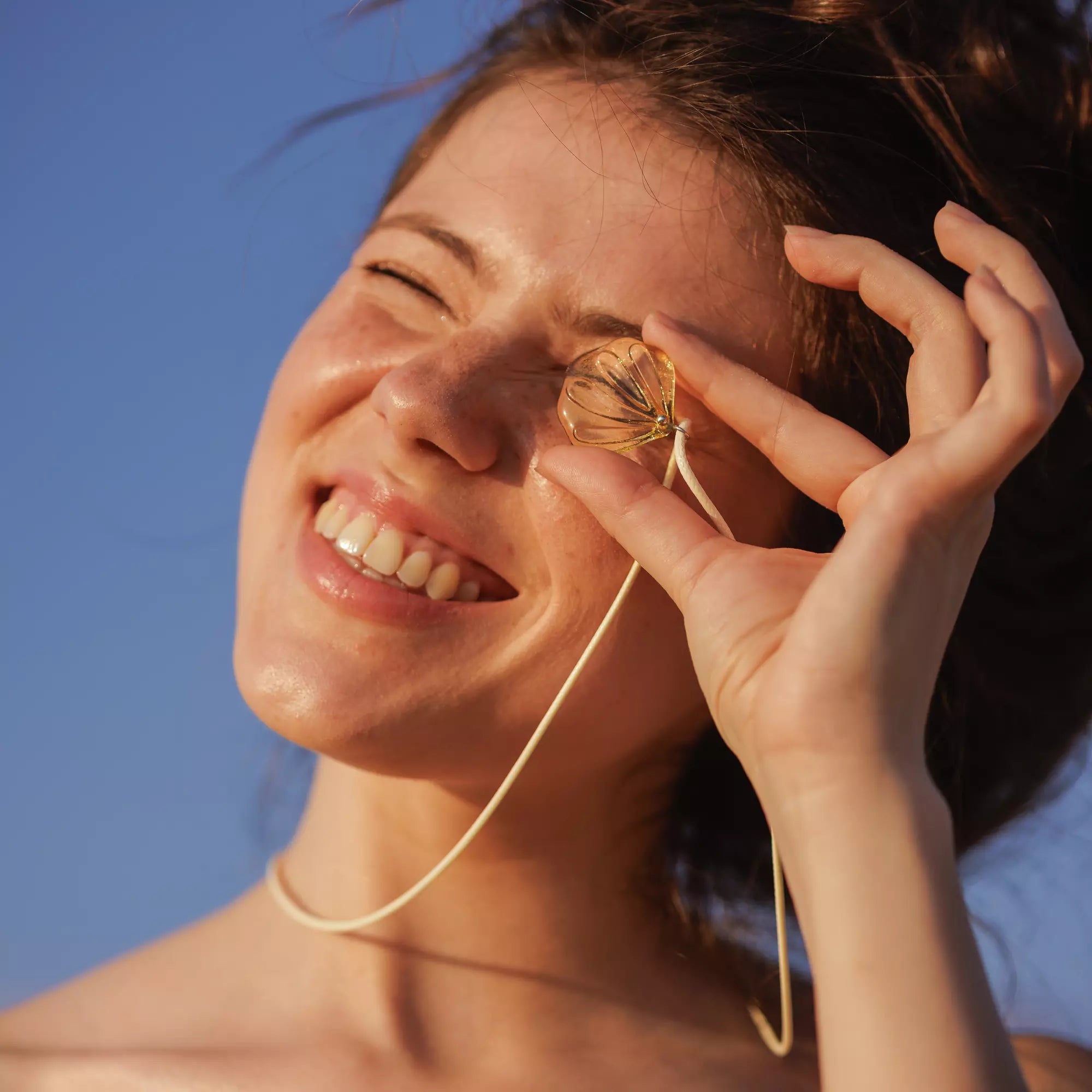 Woman holding a shell-shaped honey colored pendant near her face against a blue sky