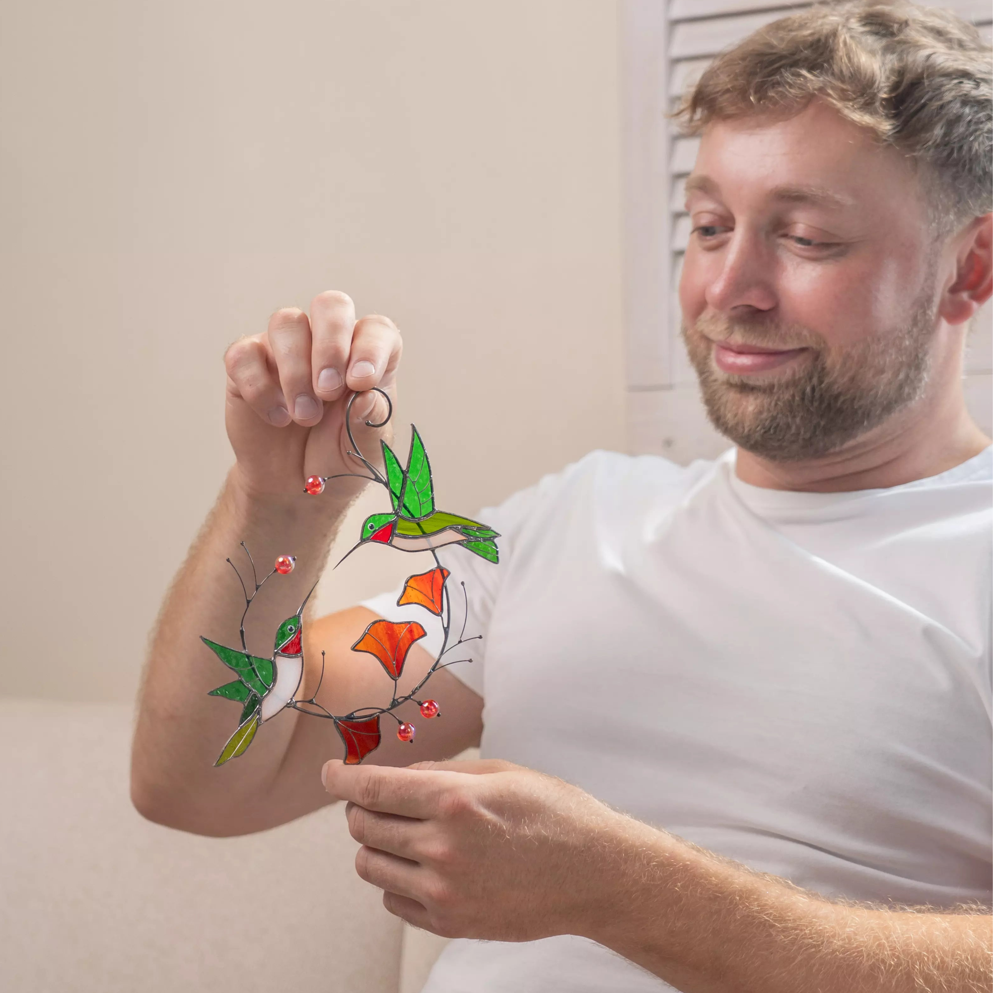 Man holding a colorful stained glass hummingbirds with flowers suncatcher against a neutral background