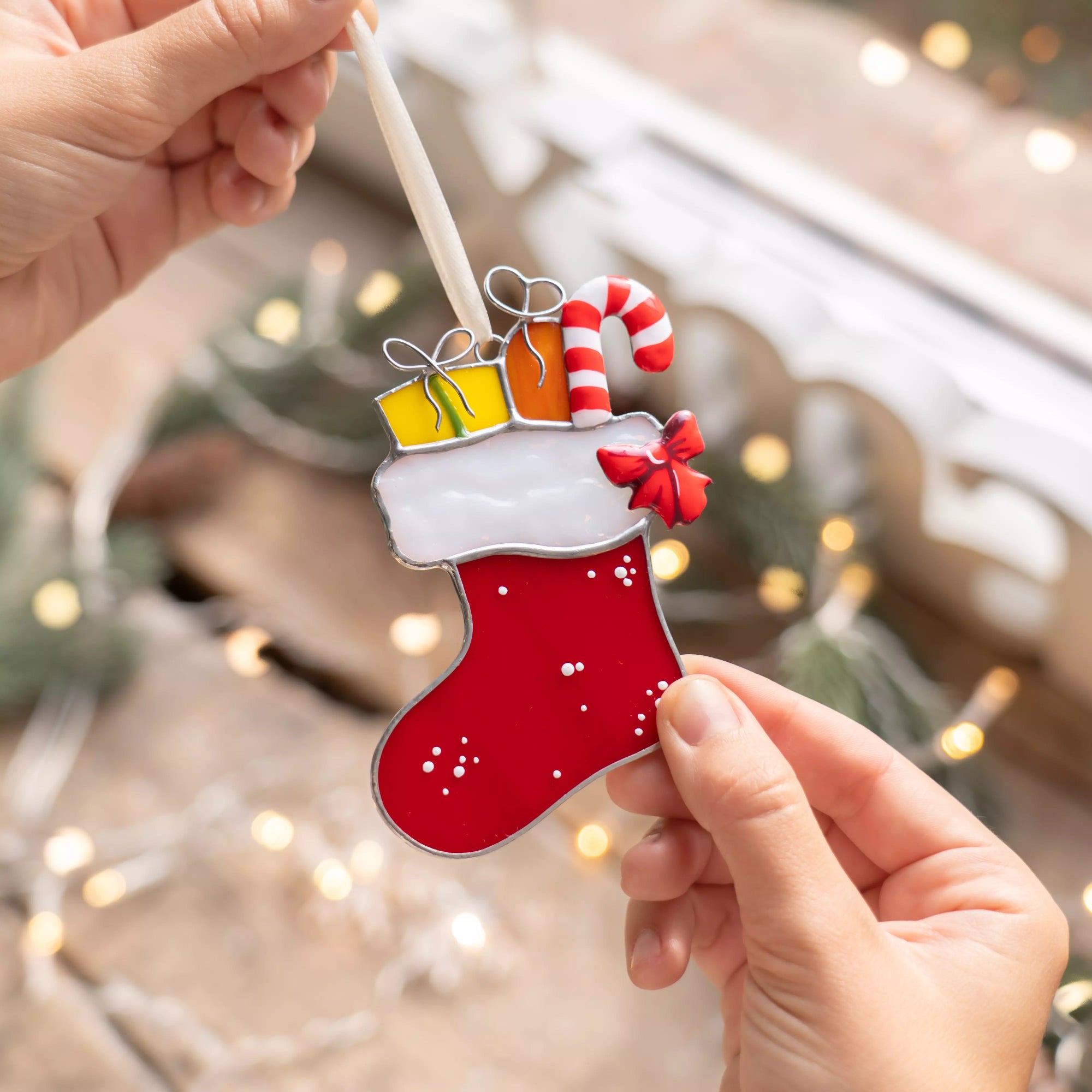 Hands holding a stained glass red Christmas stocking ornament with candy cane and yellow gifts on a wooden table.
