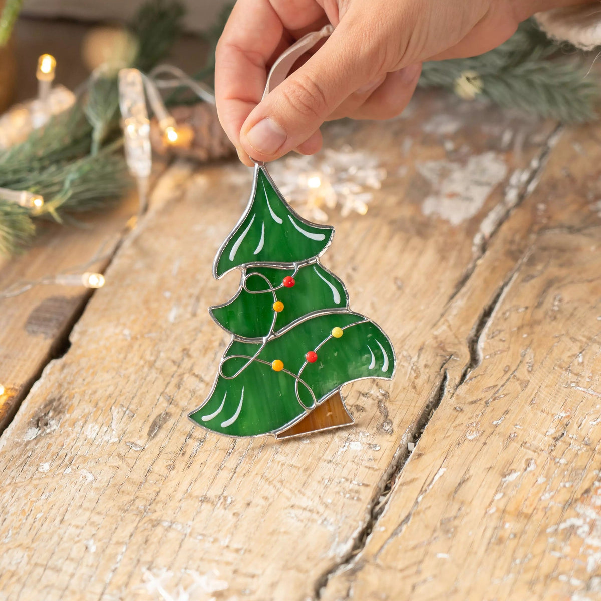 Green stained glass Christmas tree suncatcher with colorful garland placed on a rustic wooden table with fairy lights.
