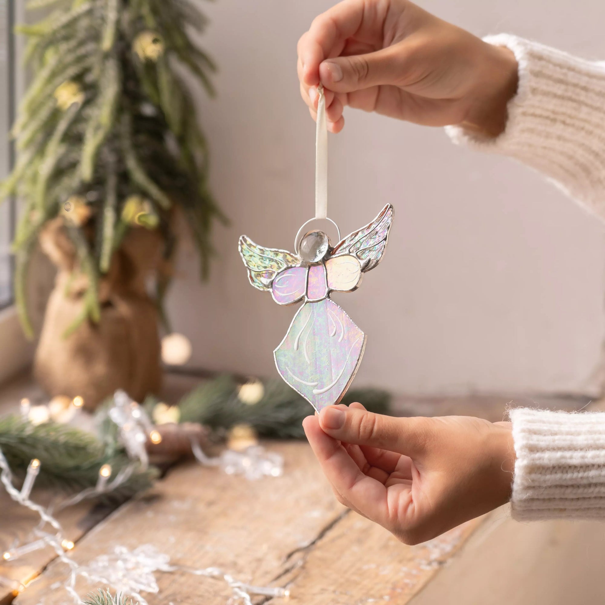 Hand holding iridescent stained glass angel above a wooden table with pine branches and fairy lights.