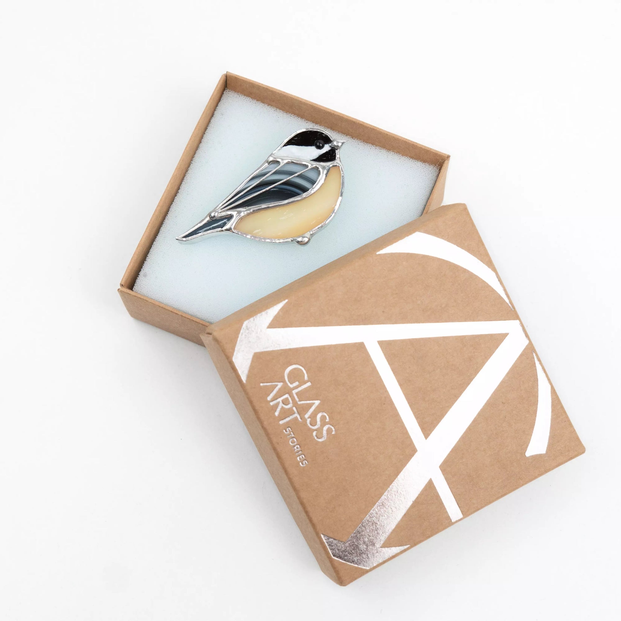 Stained glass bird brooch resting in a partially opened kraft box with Glass Art Stories logo.