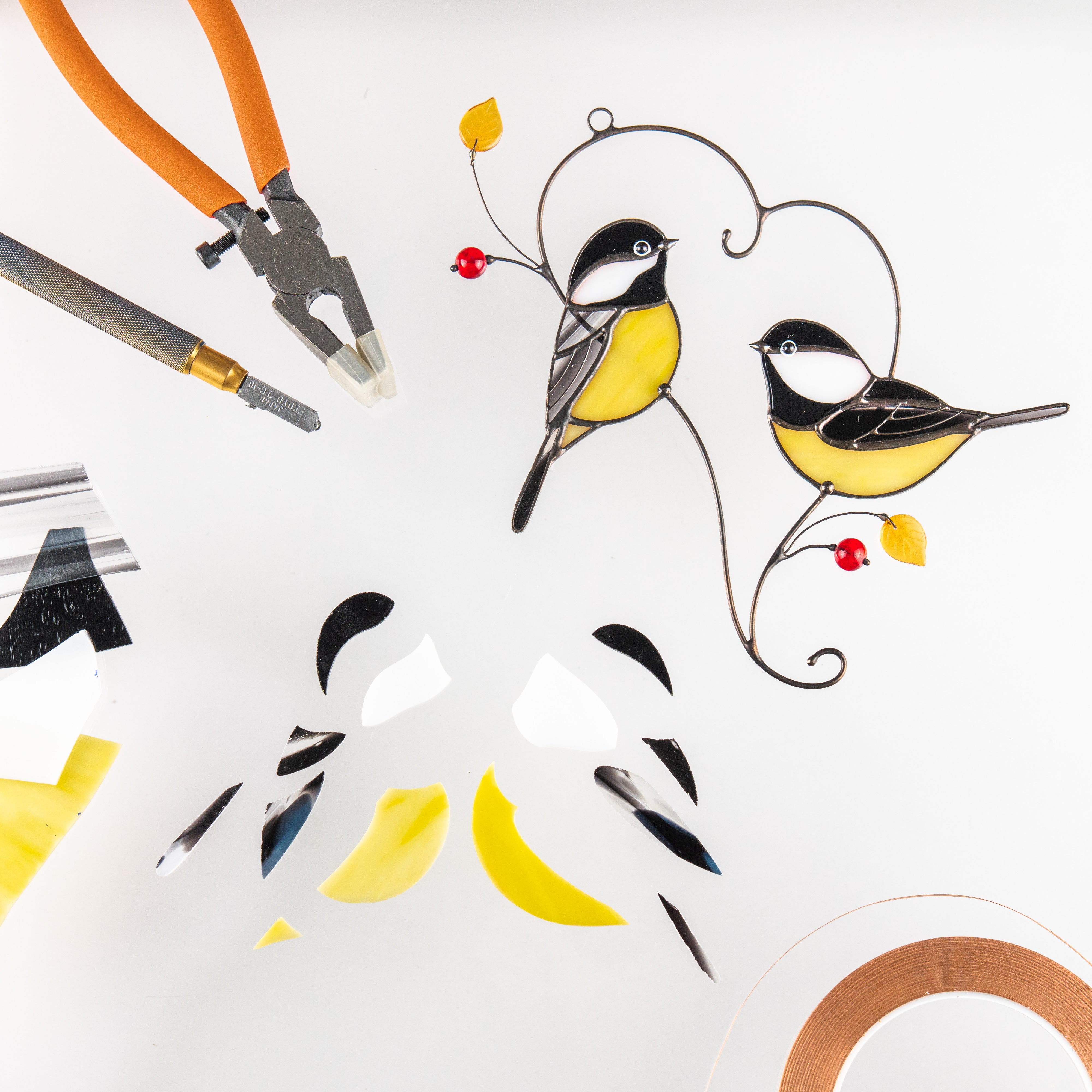 Two stained glass chickadees with yellow bellies and red beads shown alongside glass cutting tools and glass pieces.