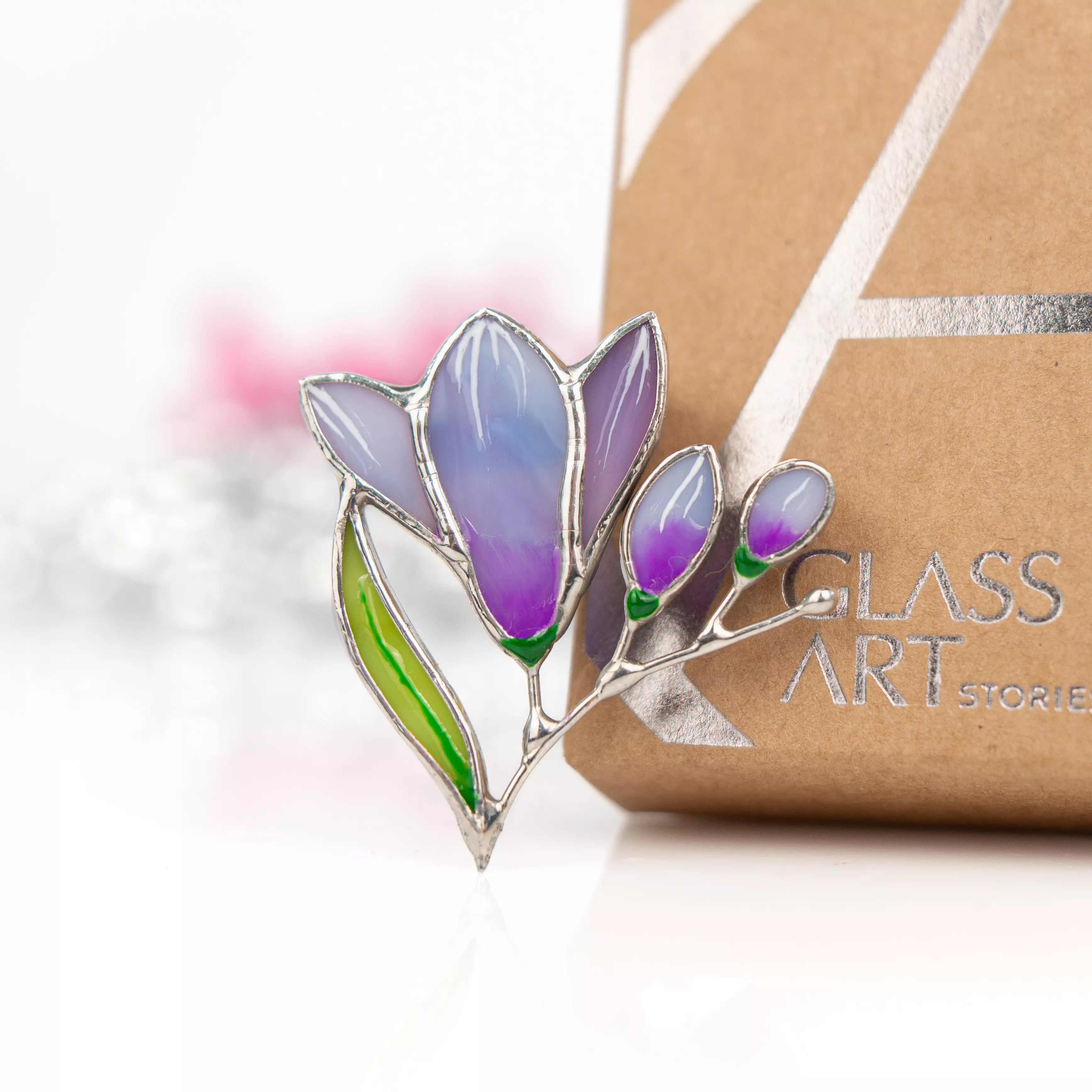 Purple stained glass freesia brooch placed near branded Glass Art Stories gift packaging