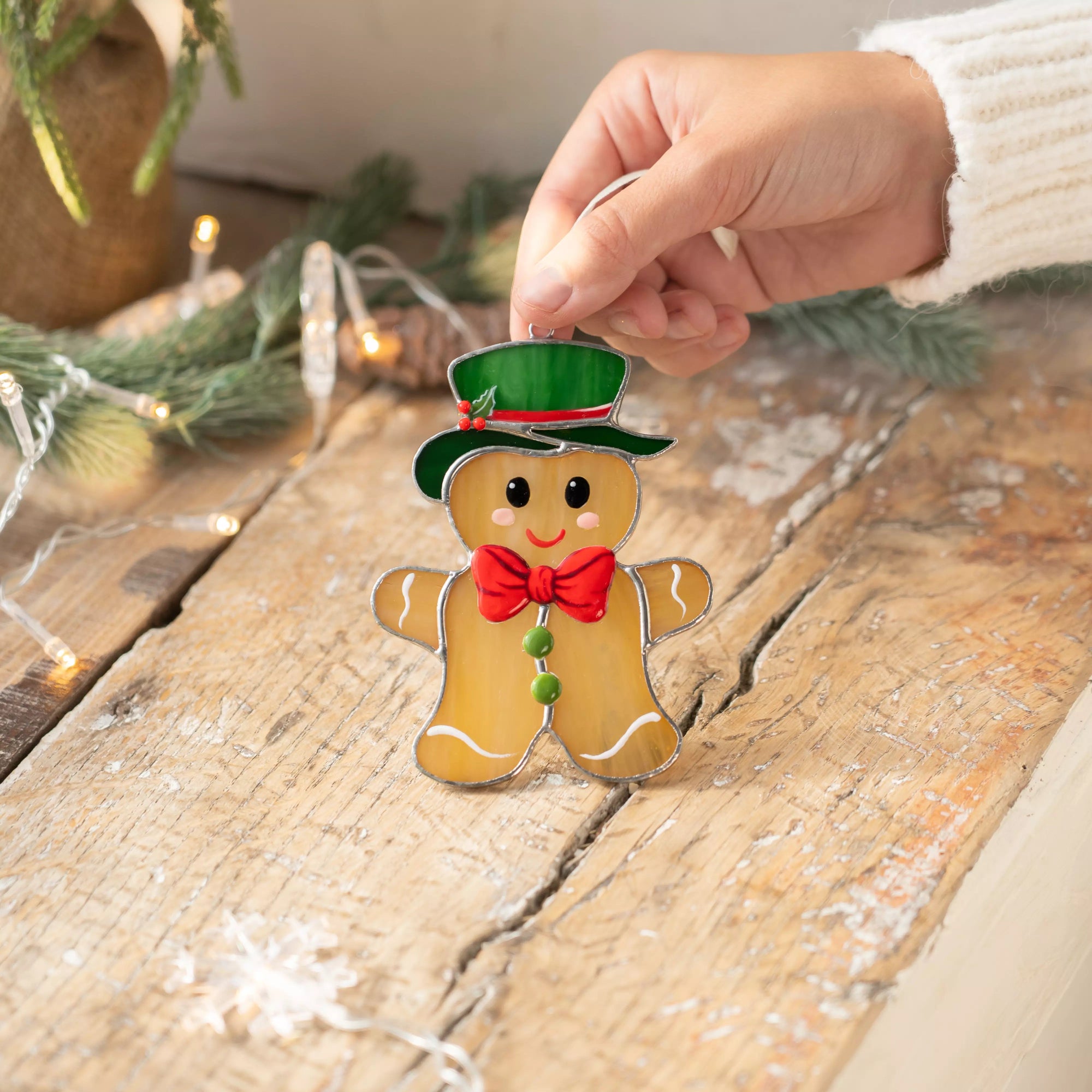 Stained glass gingerbread man with red bow and green hat lying on a rustic wooden table decorated with Christmas lights.